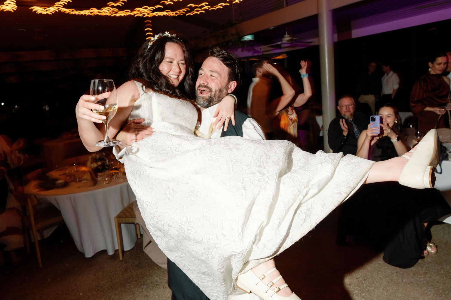 Groom lifting bride in wedding dress during lively reception dance floor celebration at night.