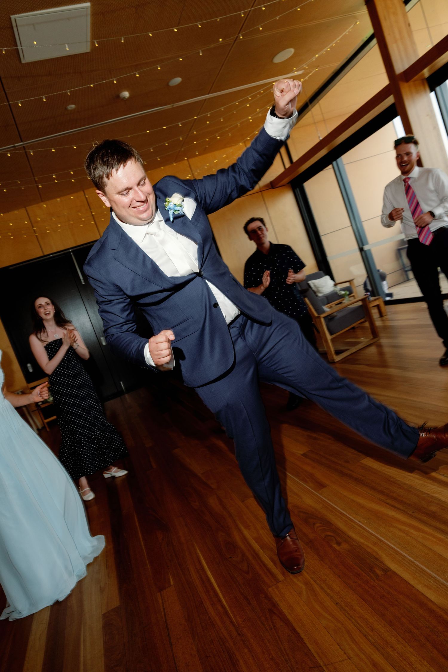 Wedding guests perform lively dance moves on hardwood floor under twinkling lights.