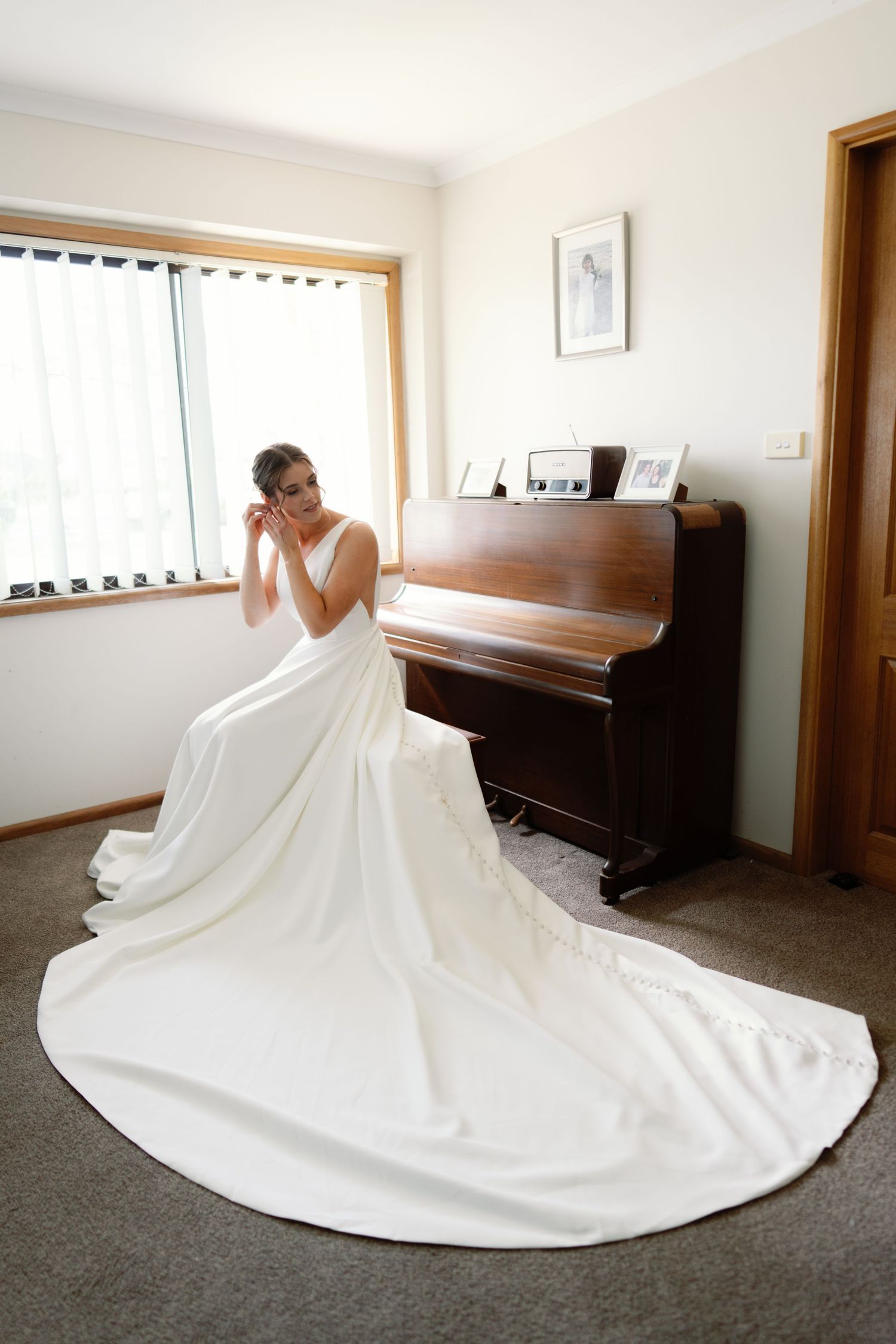 Flowing white wedding dress with long train spread out on grey carpet in sunlit room.