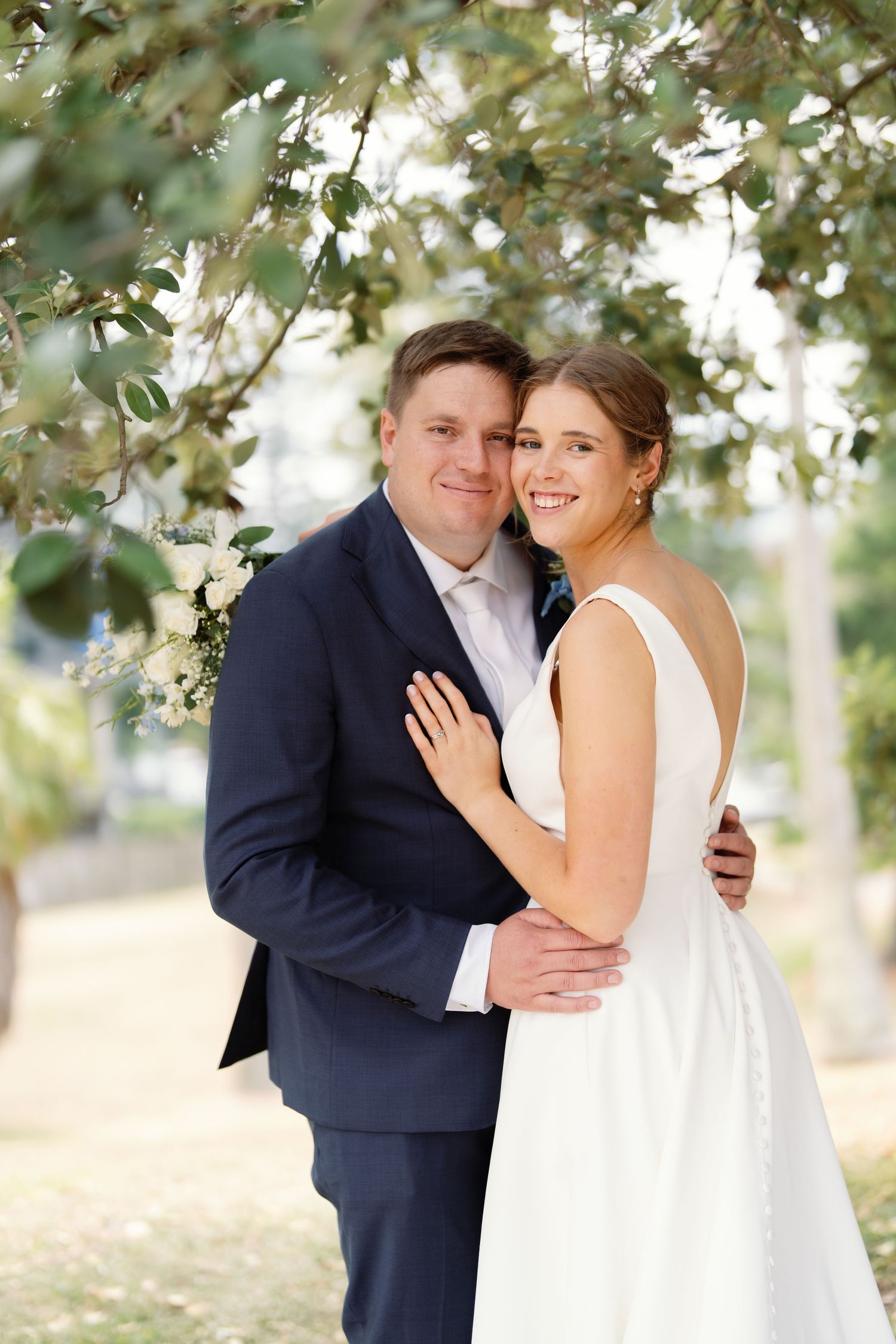 Newlyweds share an intimate moment under blooming white flowers in a garden setting.