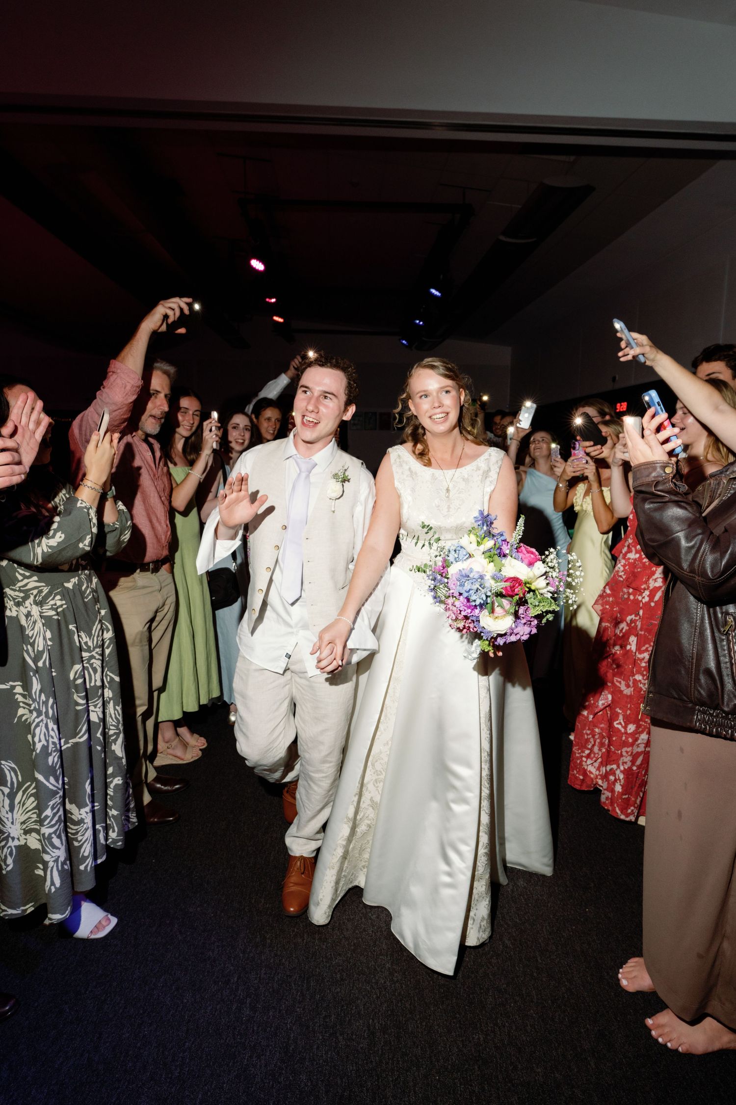 Bride and groom walk through guest sendoff line holding hands, bride carries colorful bouquet in white gown.