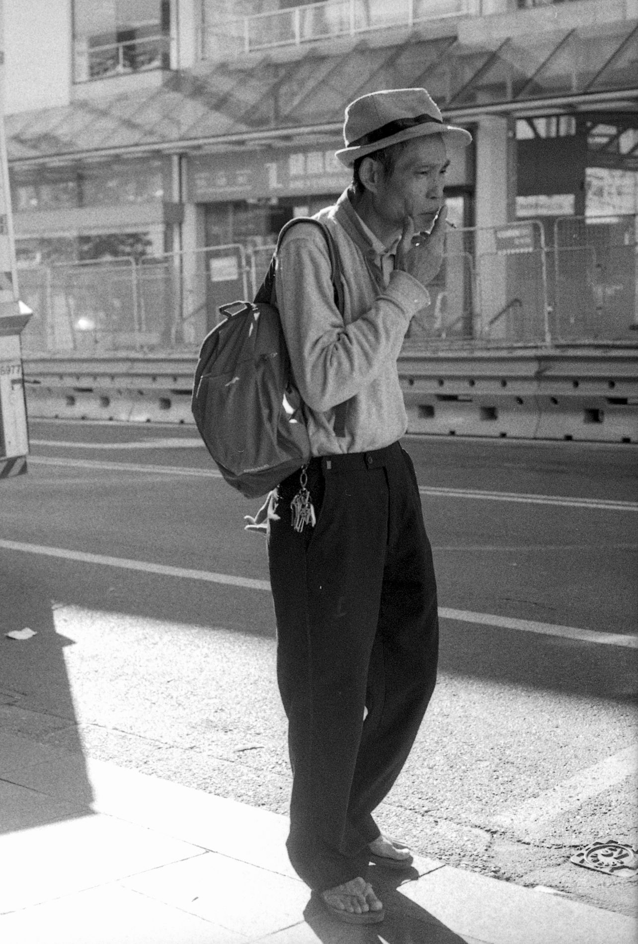 Person with backpack and hat smoking while standing on urban street in black and white photo.