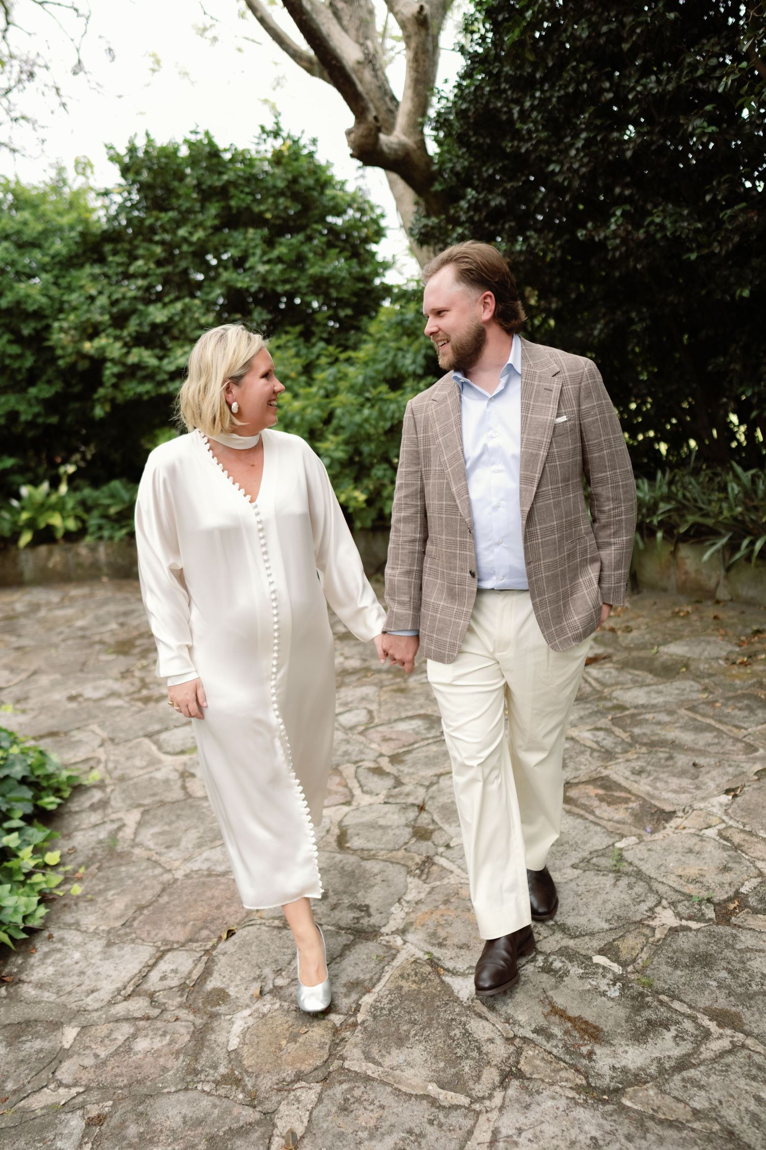A couple in coordinating cream and brown outfits walks together on a rocky path surrounded by lush greenery.