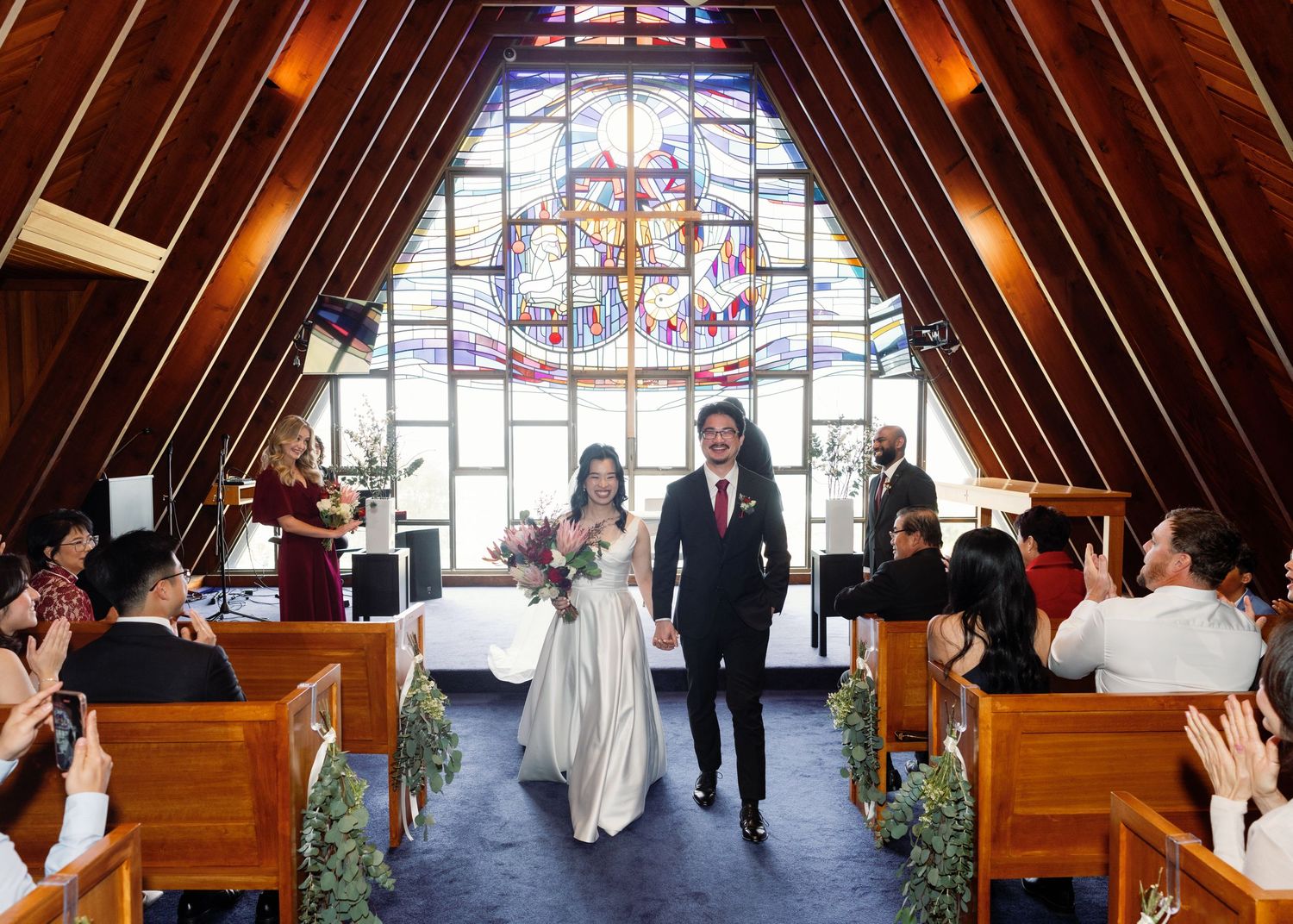 Wedding guests seated in wooden pews inside a chapel with dramatic stained glass windows and vaulted ceiling.