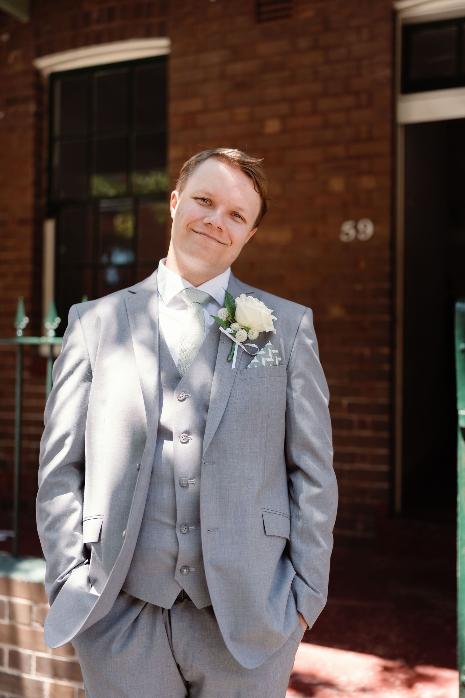 A groom in a light gray three-piece suit poses against a brick building on his wedding day.