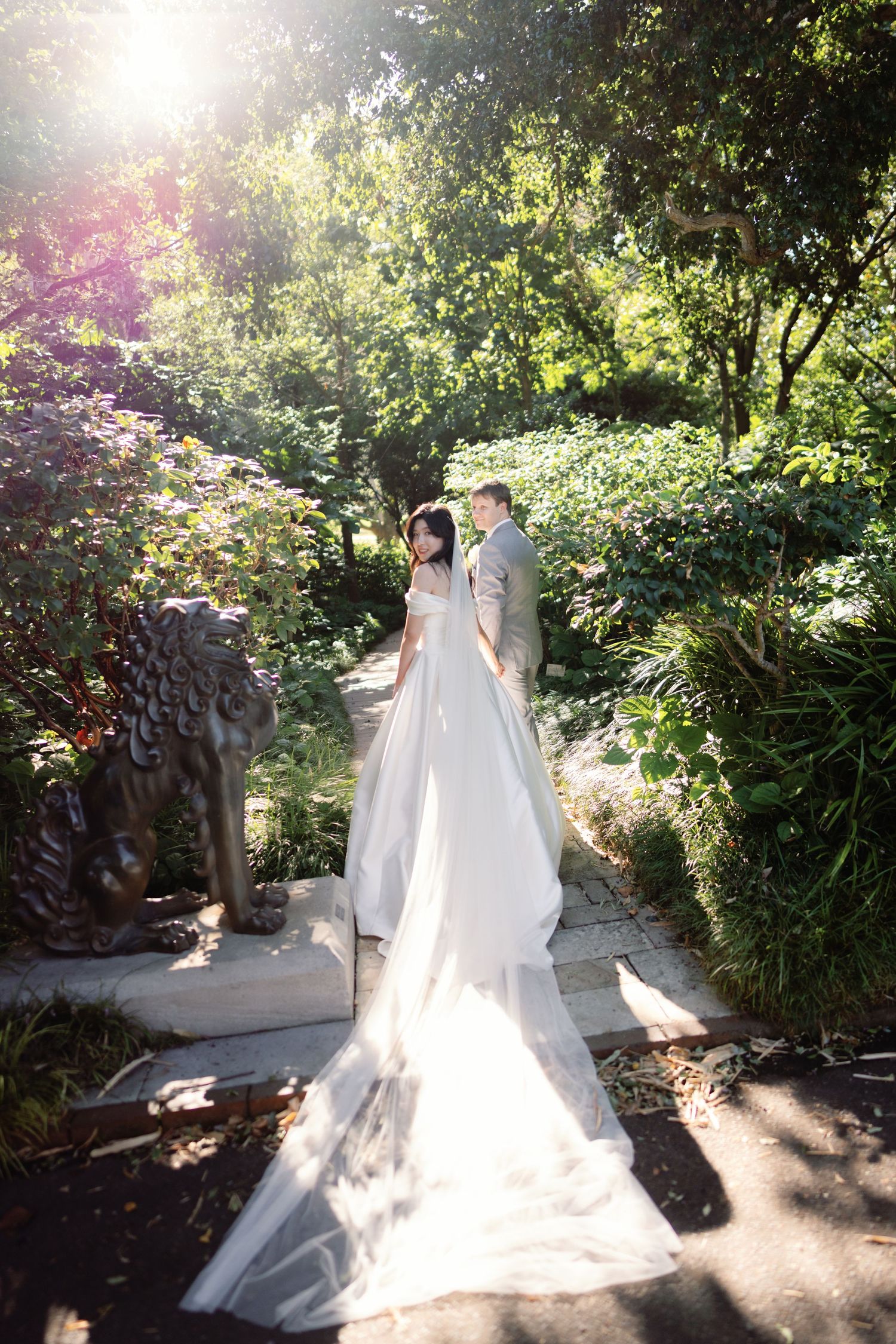 Bride in elegant white gown with long train stands with groom in lush garden, sunlight filtering through trees.