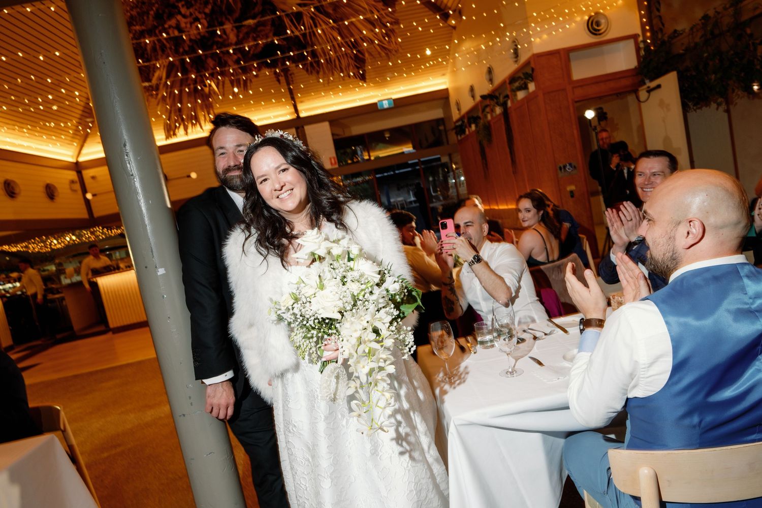 Bride in white fur wrap holding floral bouquet walks with groom through cheering wedding reception guests.
