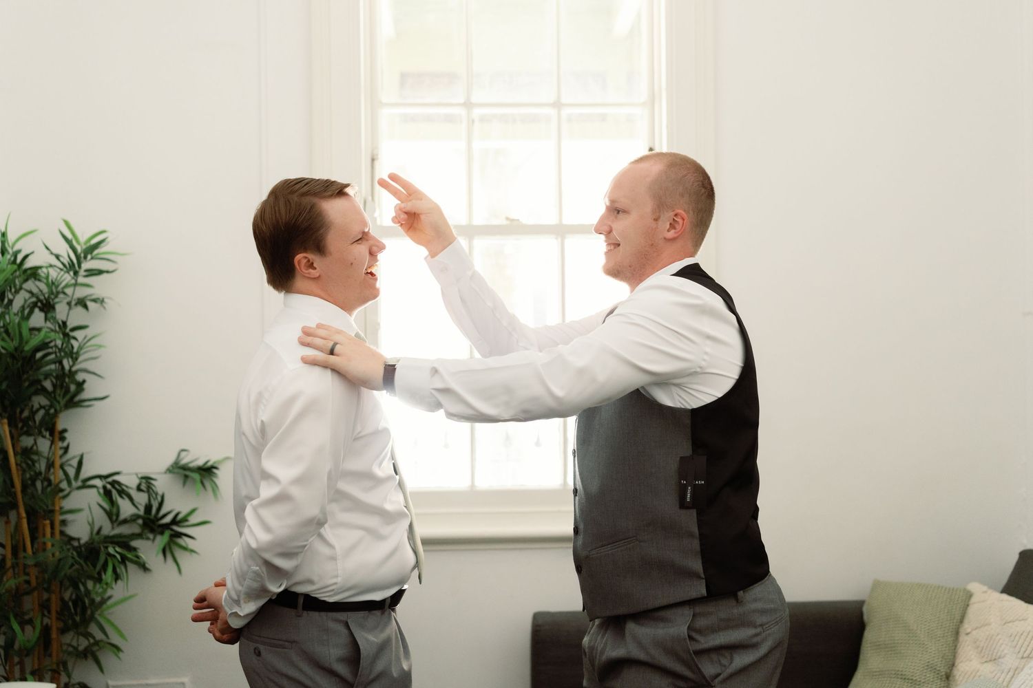 Two people in formal attire appear to be in a playful confrontation by a window in a bright room.