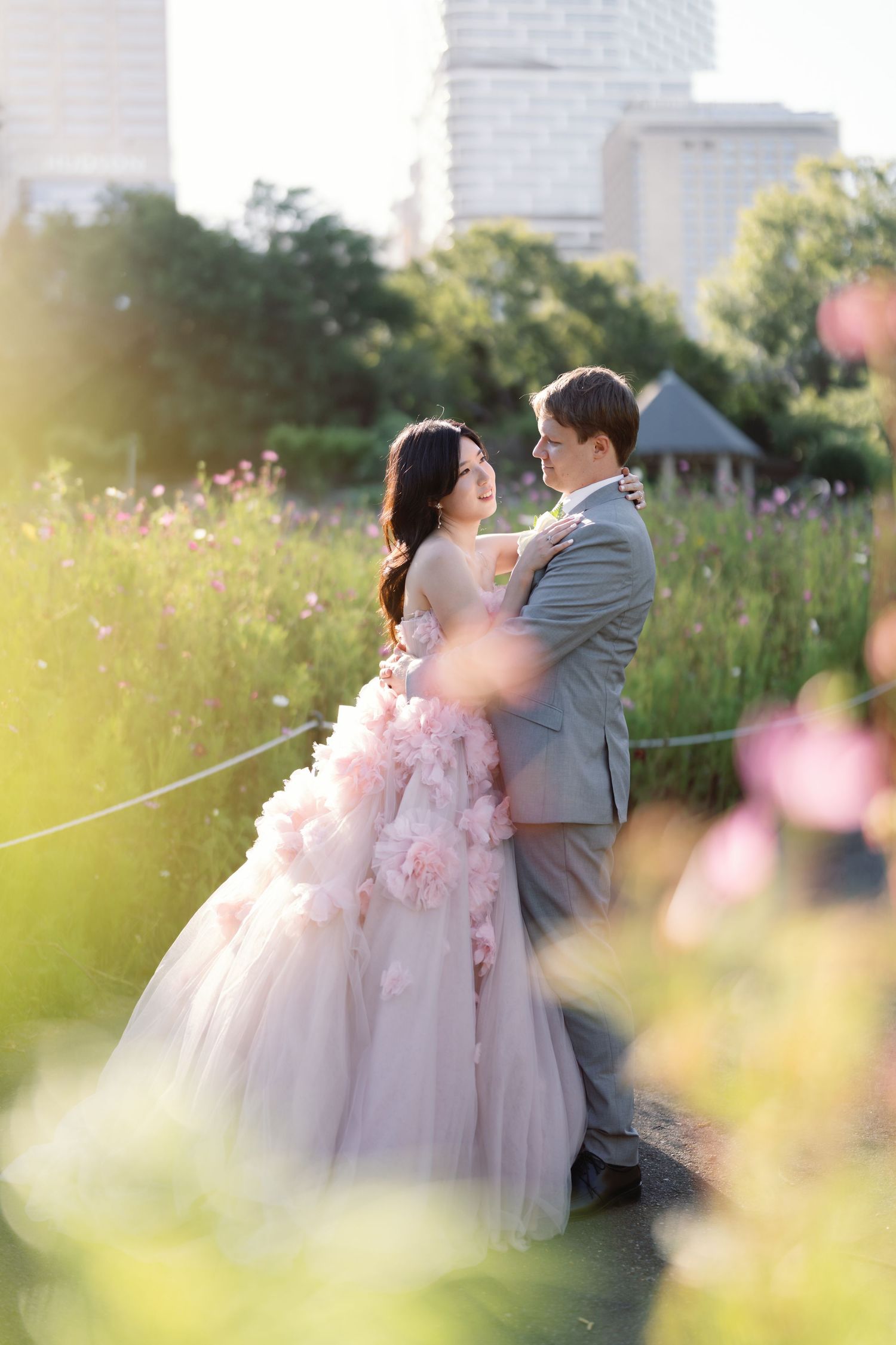 Romantic wedding couple embracing in a sunlit wildflower field with city skyline in background.