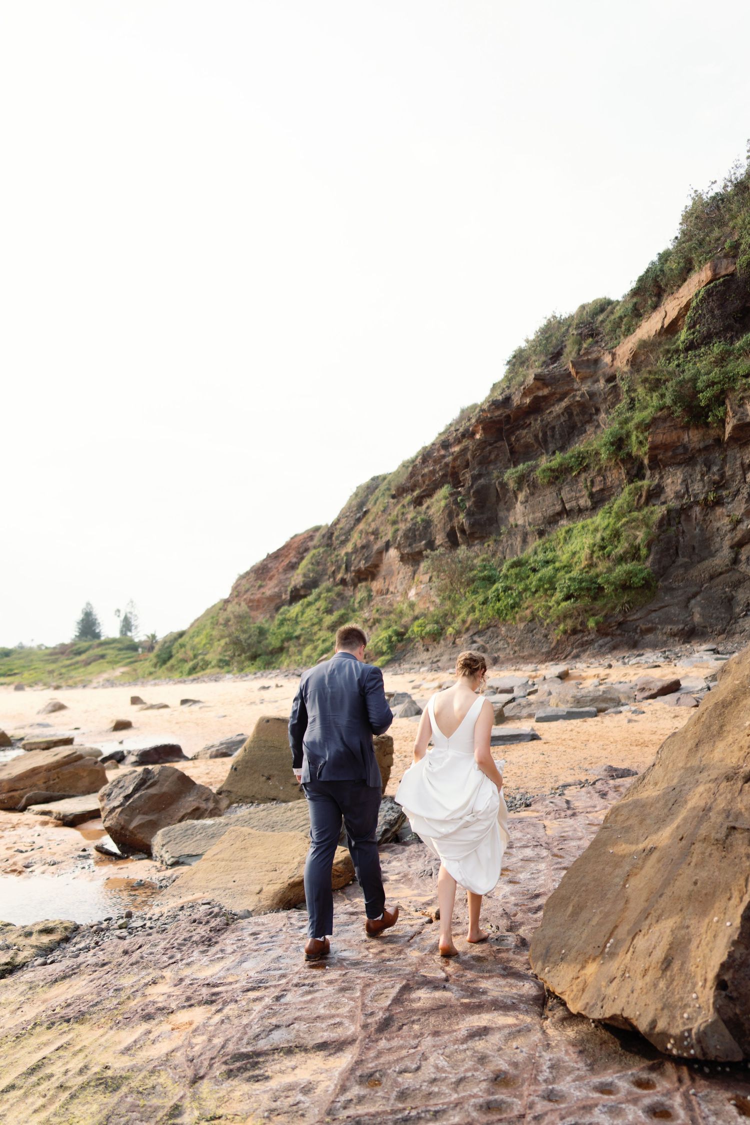A couple walking along a rocky beach coastline during a wedding photoshoot.