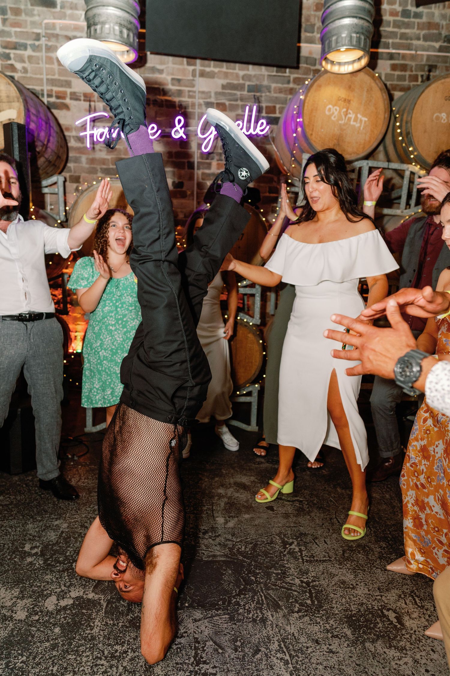 Wedding party dances energetically at a reception venue with exposed brick walls and ambient purple lighting.