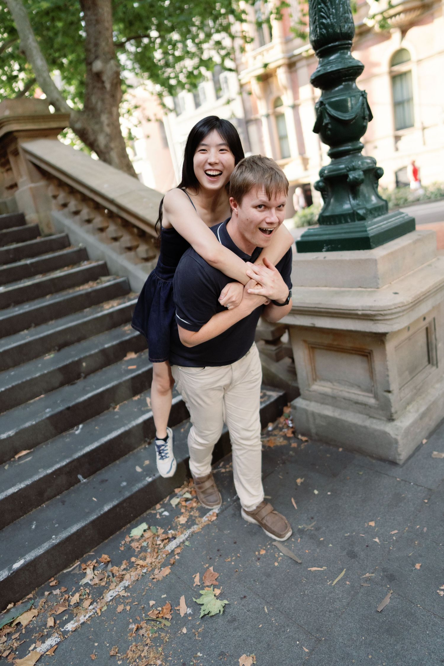 A playful couple shares a piggyback ride moment on urban steps near an ornate green lamppost.