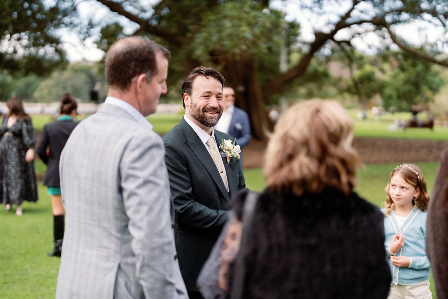 Groom in dark suit laughing with wedding guests on green lawn during outdoor wedding reception.
