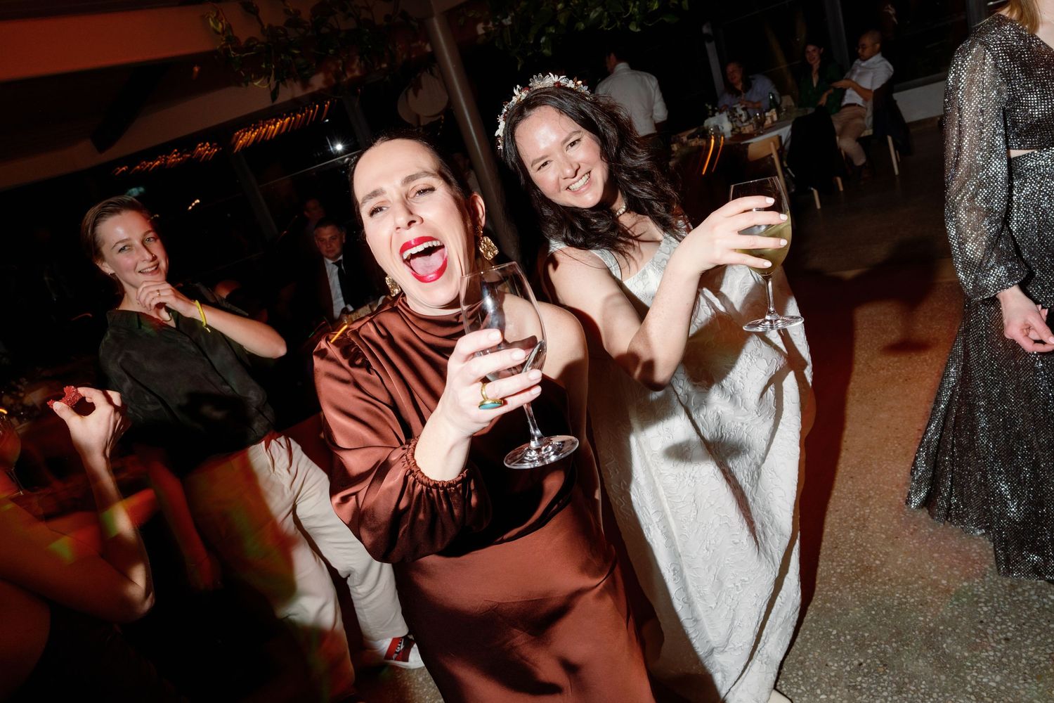 Two women in elegant dresses laughing and holding champagne glasses at an outdoor wedding reception.