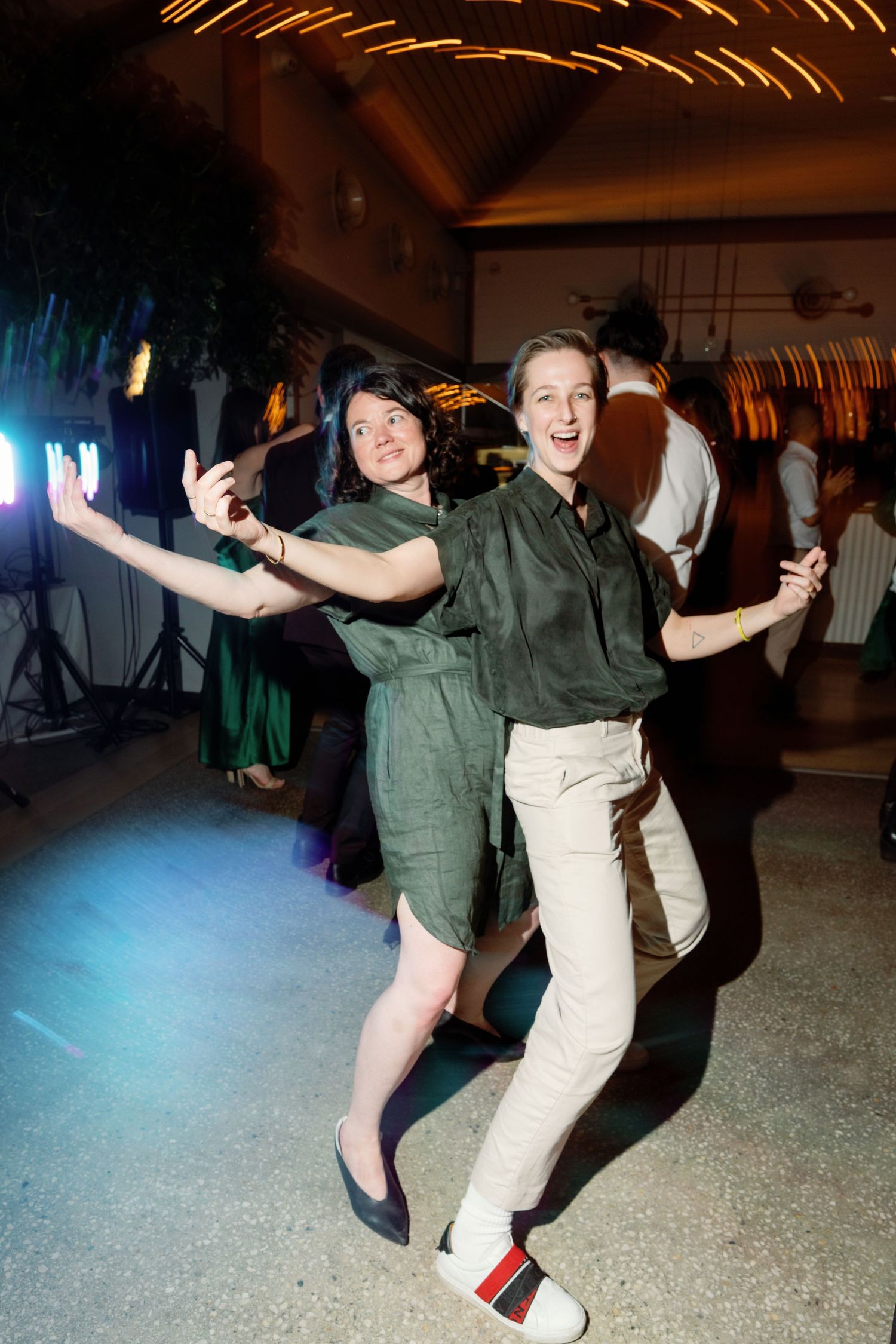 Two women in green outfits dancing joyfully at an indoor party event with colorful lights.