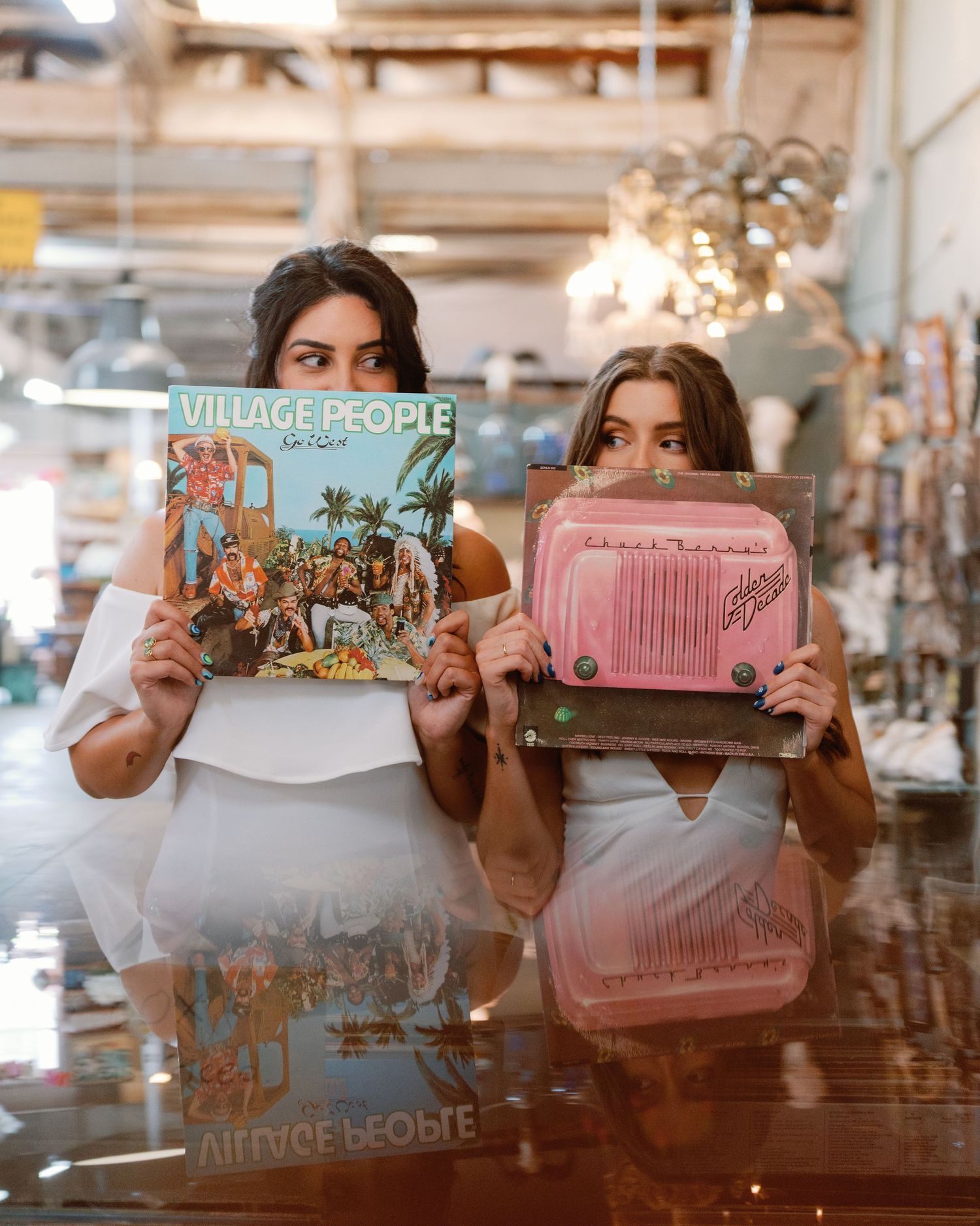 People holding up vintage vinyl records in a reflective surface at a retro-styled shop interior.