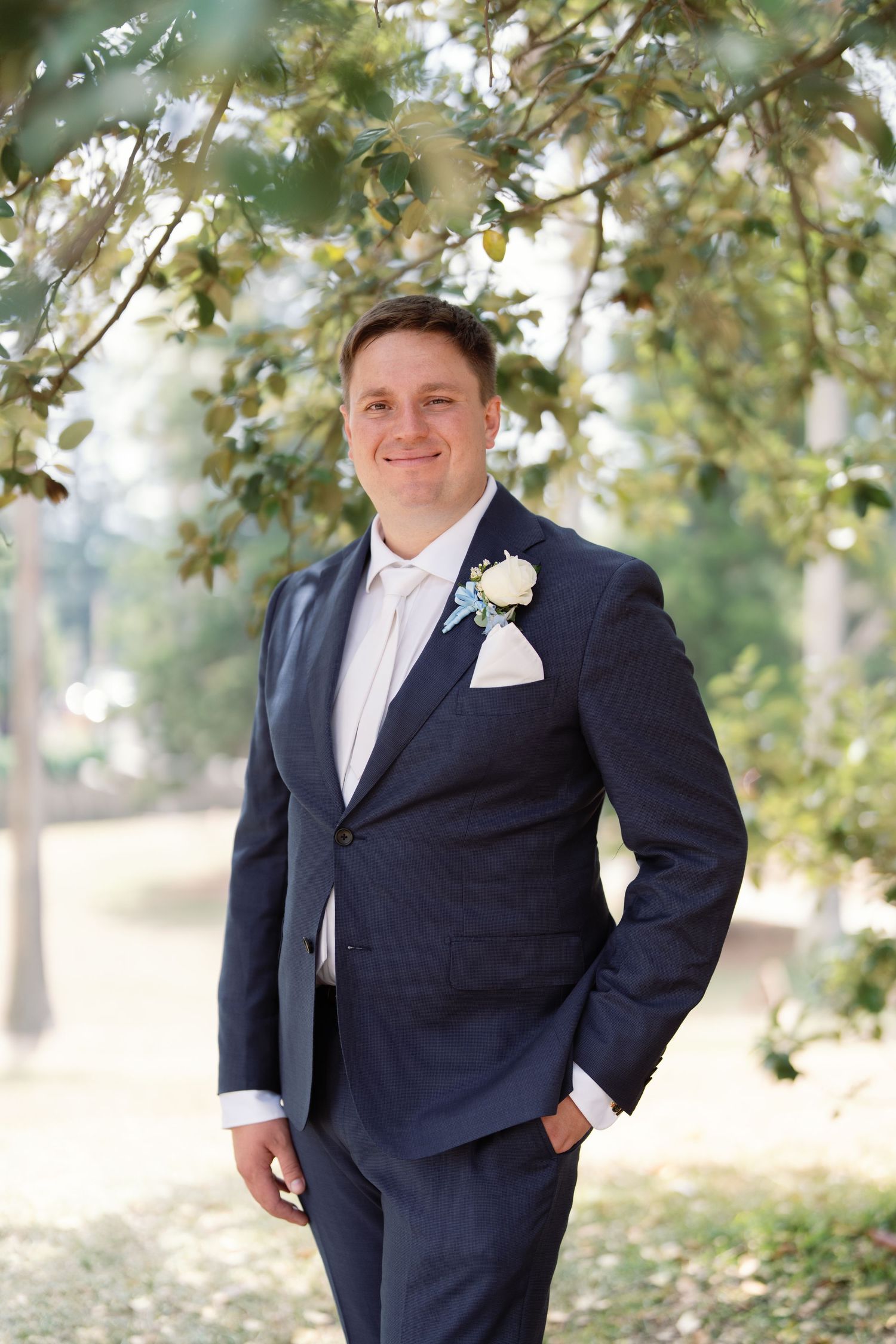 Groom in navy suit with white boutonniere poses under green leafy trees.