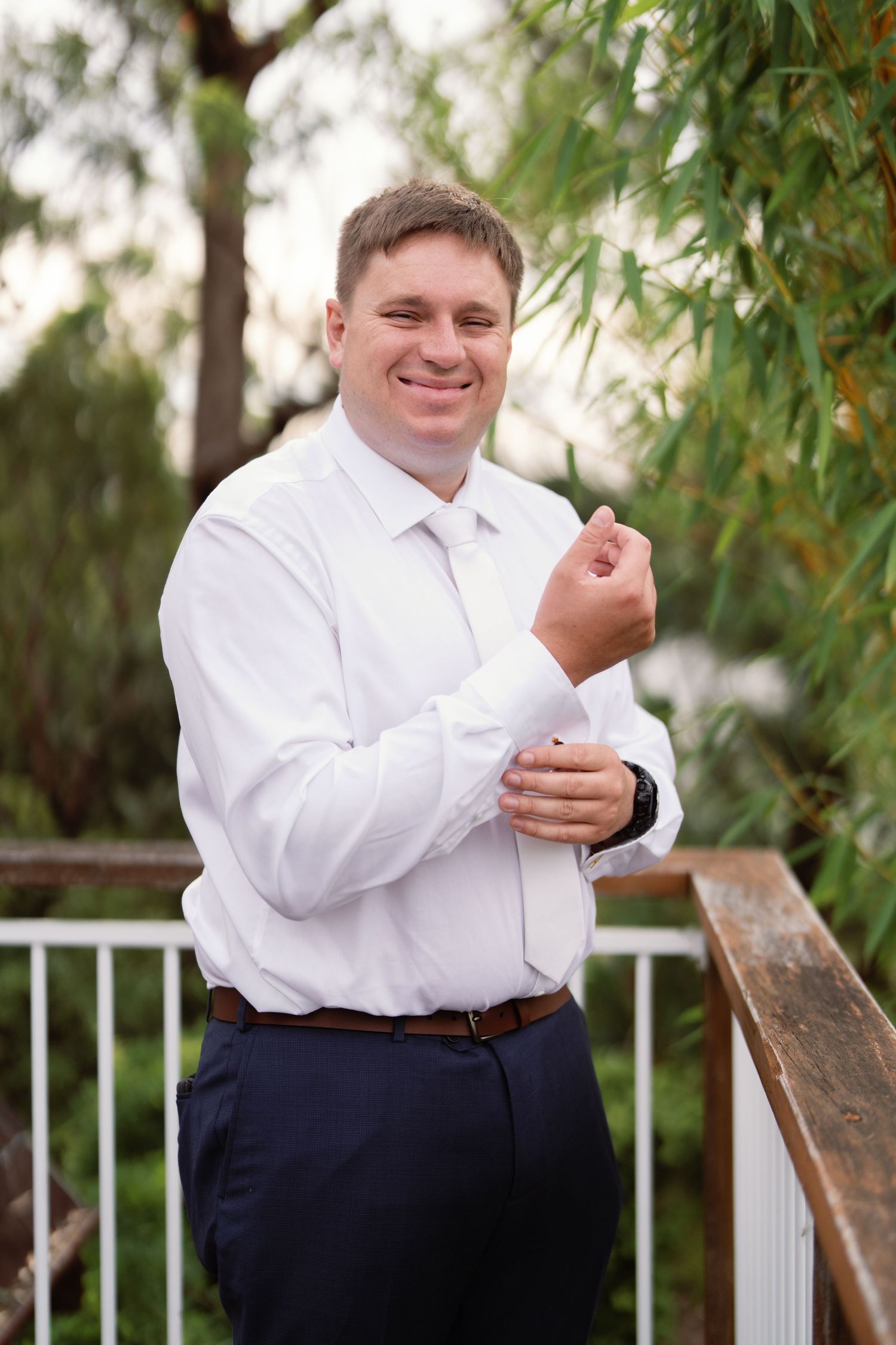 A groom in a white dress shirt and dark pants smiles while standing outdoors near foliage.