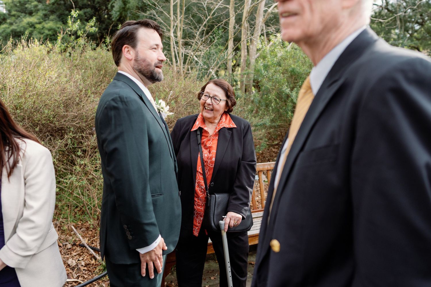A group of people in formal attire gather outdoors, smiling and conversing near trees at what appears to be a wedding.
