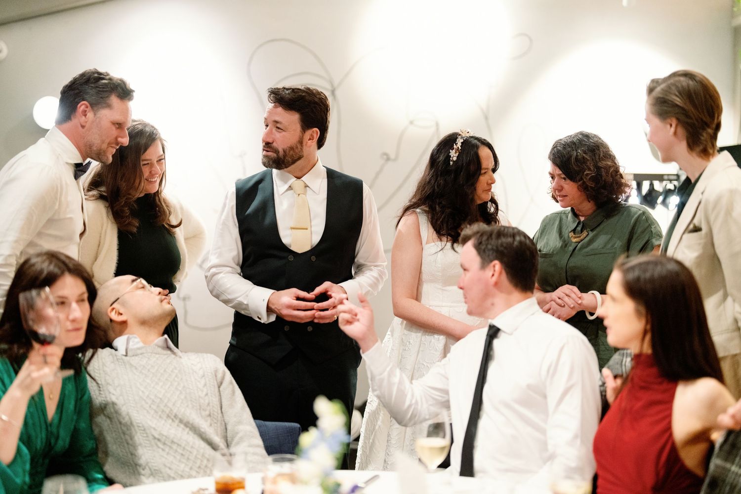 A bride and groom in a black vest interact warmly with wedding guests at an indoor reception gathering.