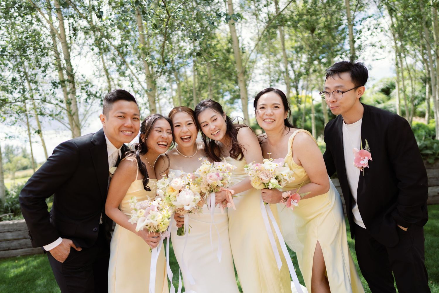 Wedding party in formal attire poses outdoors among birch trees on sunny day.