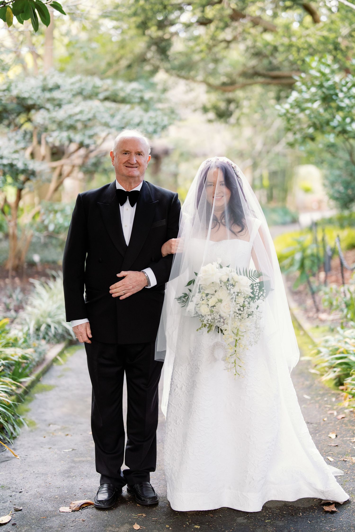 Bride in white lace gown and veil walks with older man in tuxedo through lush garden path.
