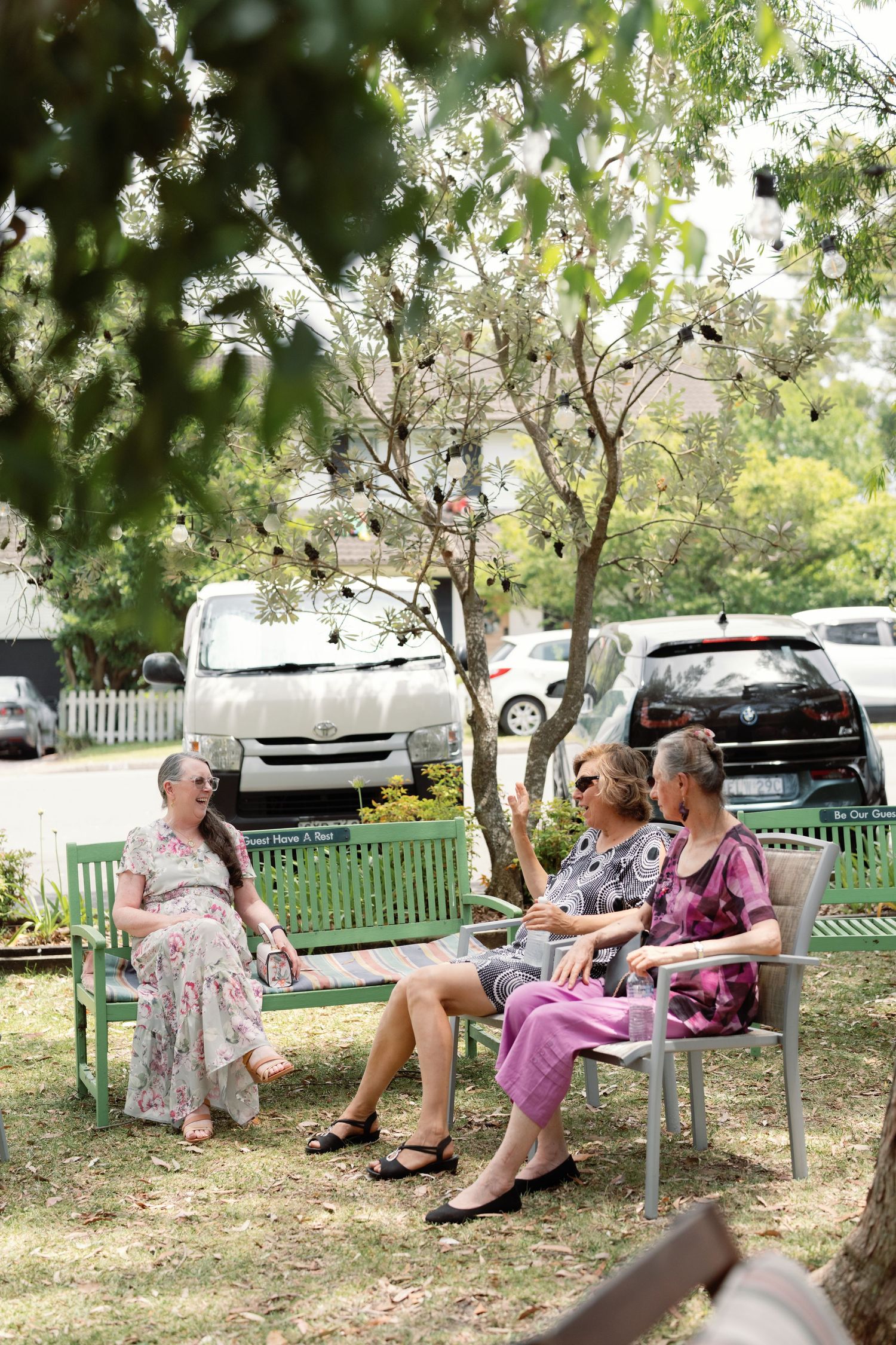 Wedding guests relax on green benches in a shaded outdoor garden area.