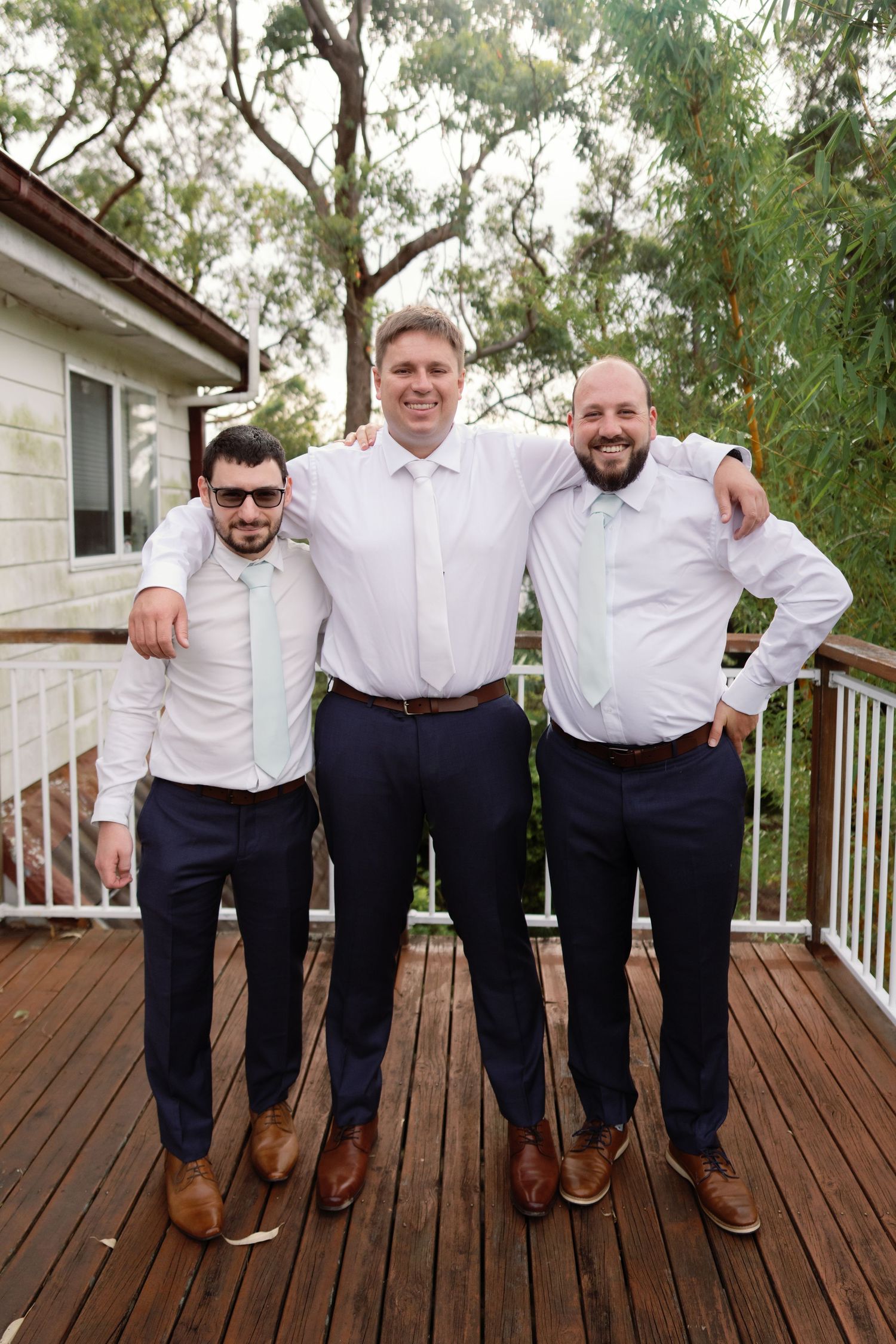Groomsmen in matching white shirts and navy pants pose playfully on a wooden deck.