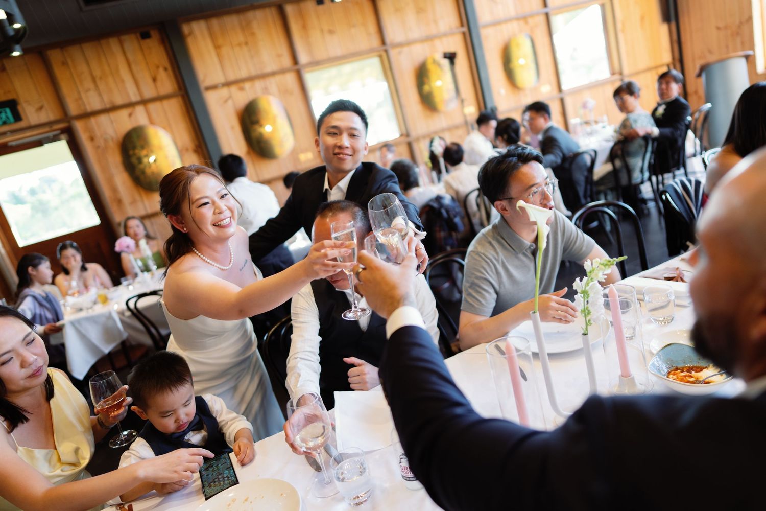 Wedding reception guests enjoying a meal and conversation at tables in a wooden-paneled venue.