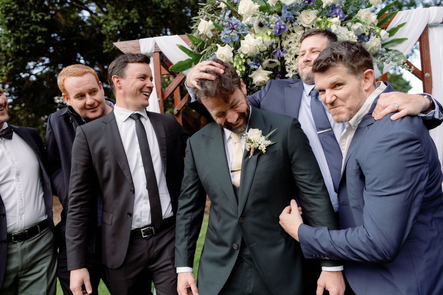 Groom in green suit laughing with groomsmen in blue suits at outdoor wedding ceremony with floral arch.