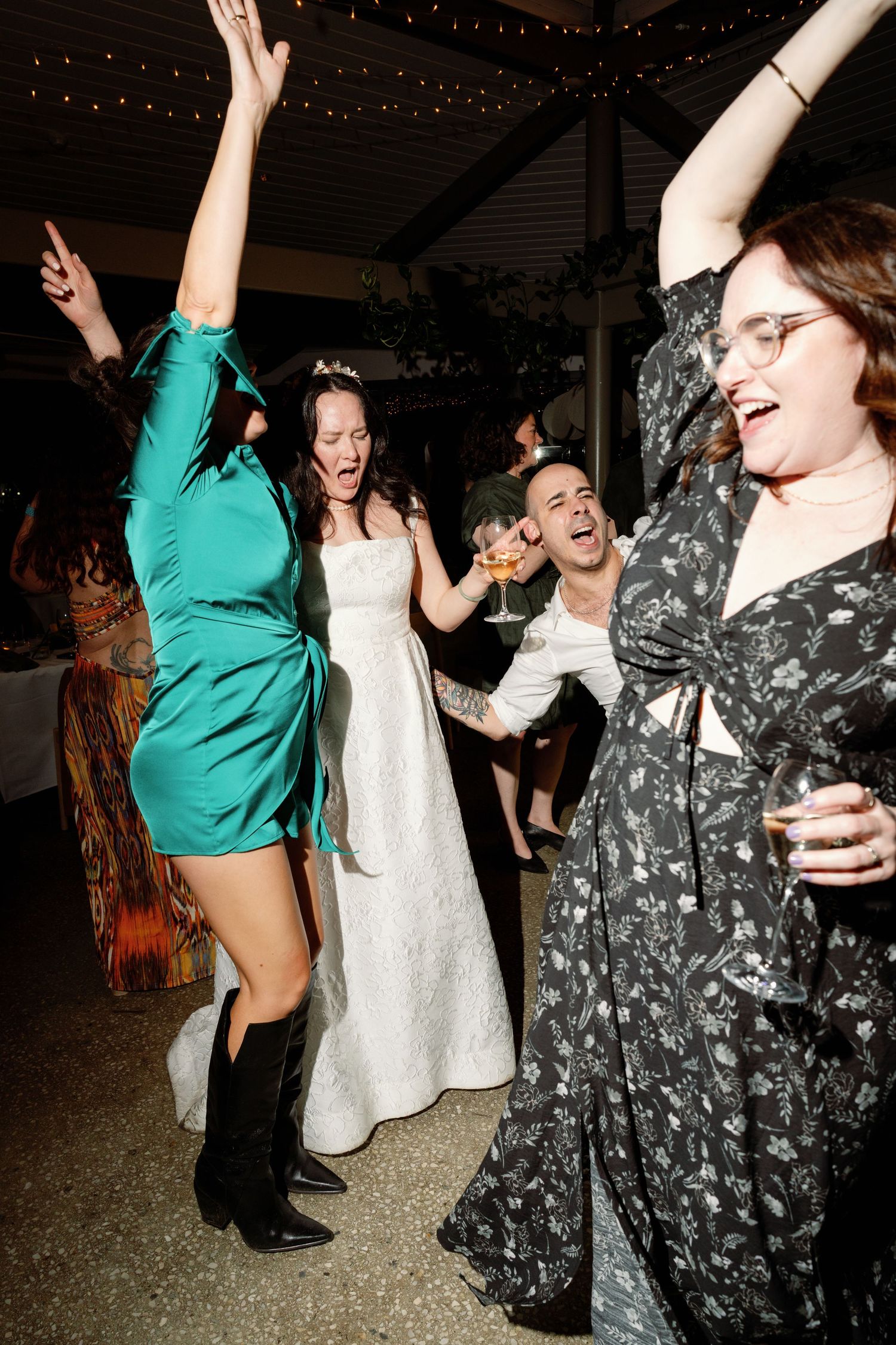 Bride in white gown dances joyfully with guests in green dress and floral outfit at lively wedding reception.