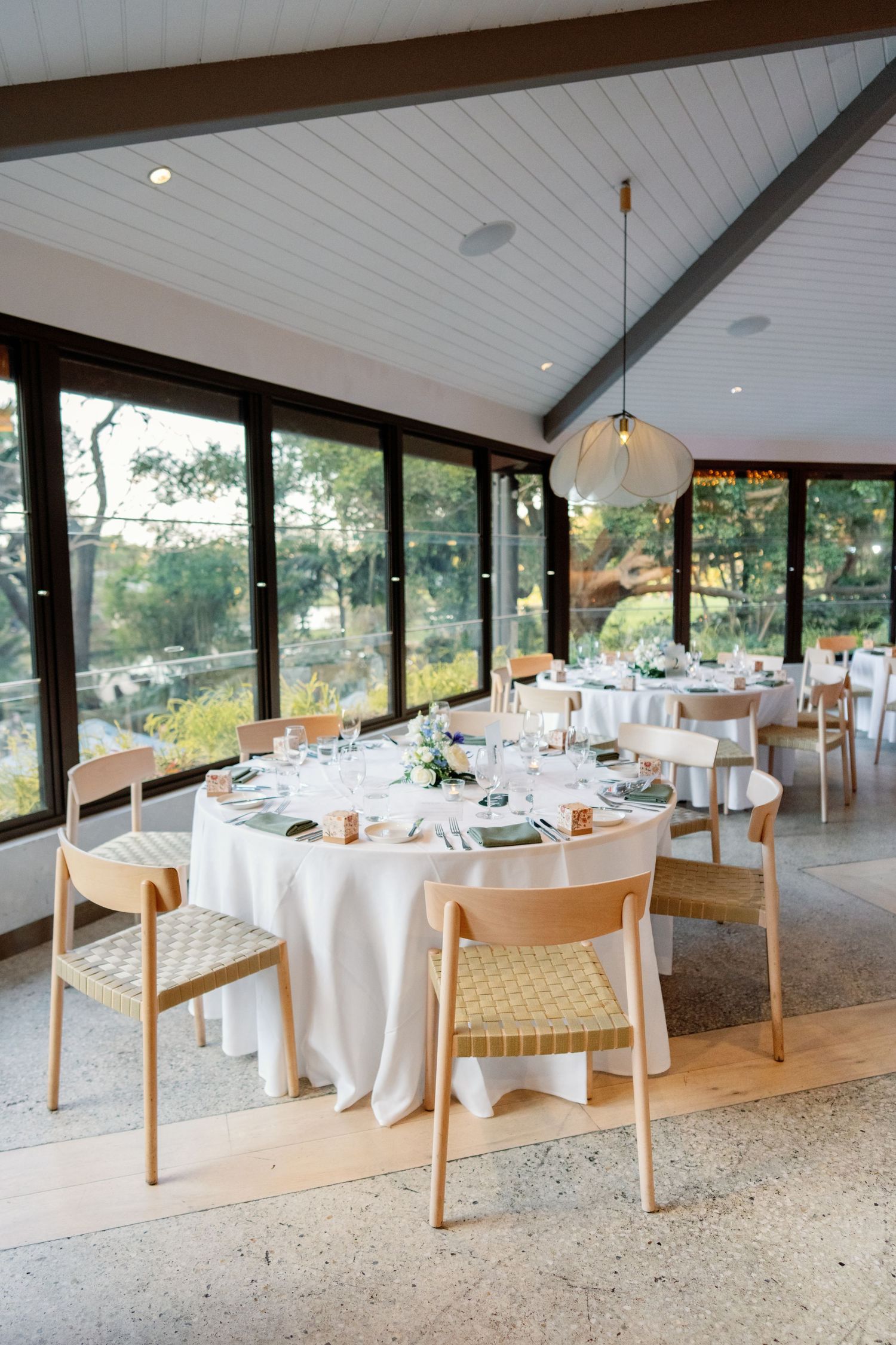 Elegant wedding reception venue with round tables, white linens, wooden chairs, and large windows overlooking lush greenery.