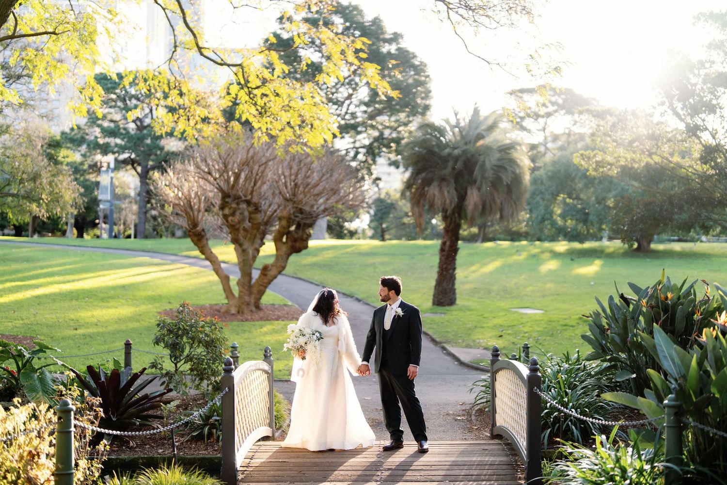 Bride and groom walk hand-in-hand across a wooden bridge in a lush garden at golden hour.