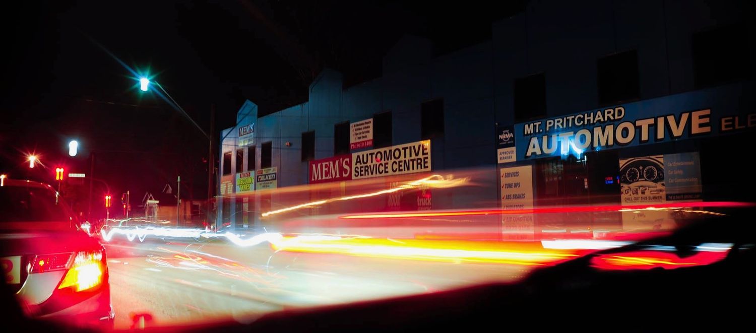 Light trails from cars streak past storefronts on a dark city street.