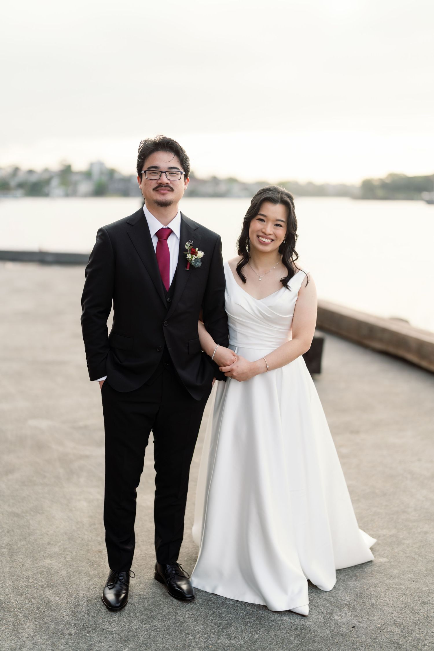 A couple in formal wedding attire poses together on a waterfront walkway.