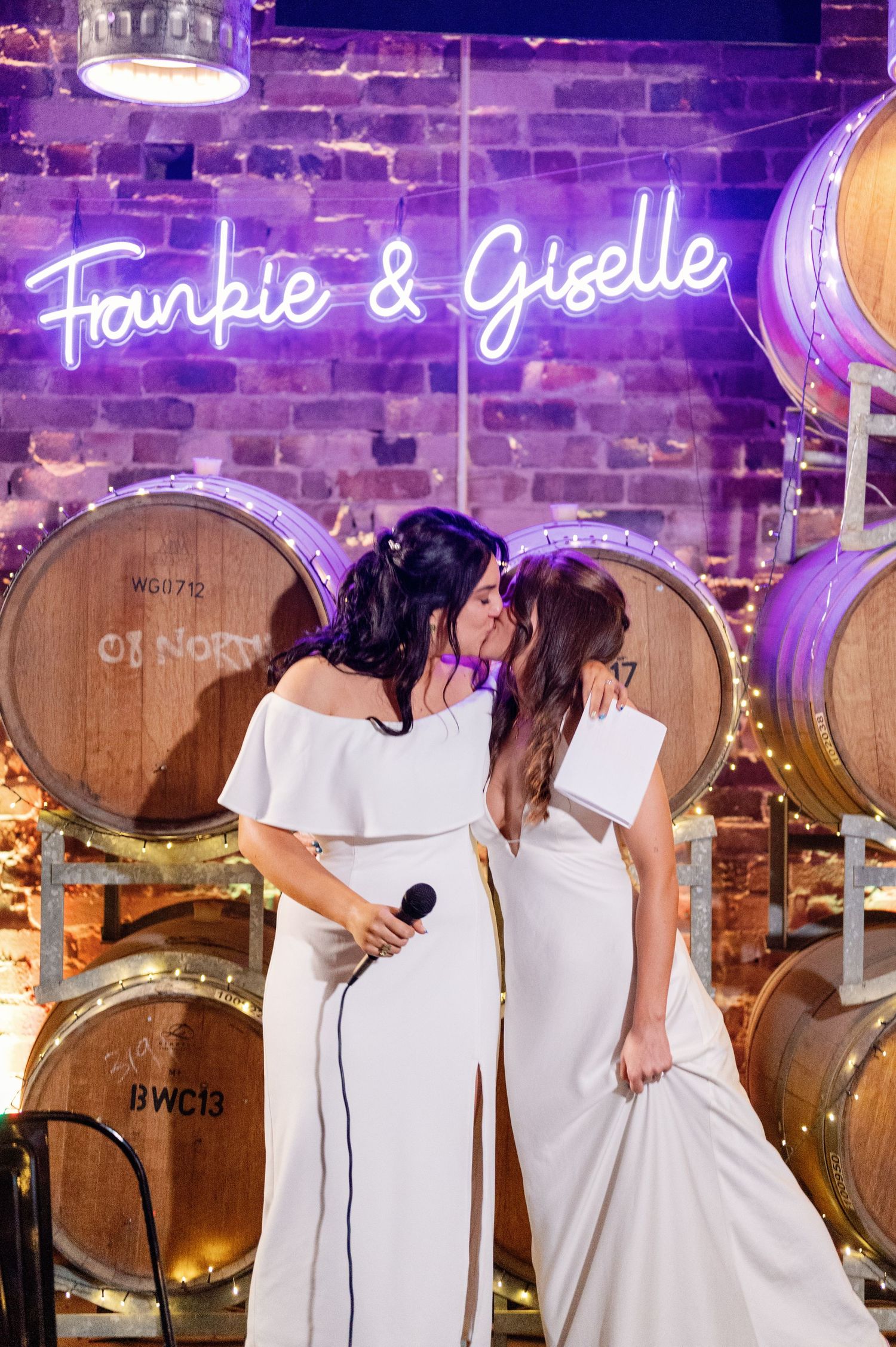Friends in matching white outfits embrace in a wine cellar with purple neon signs and wooden barrels.