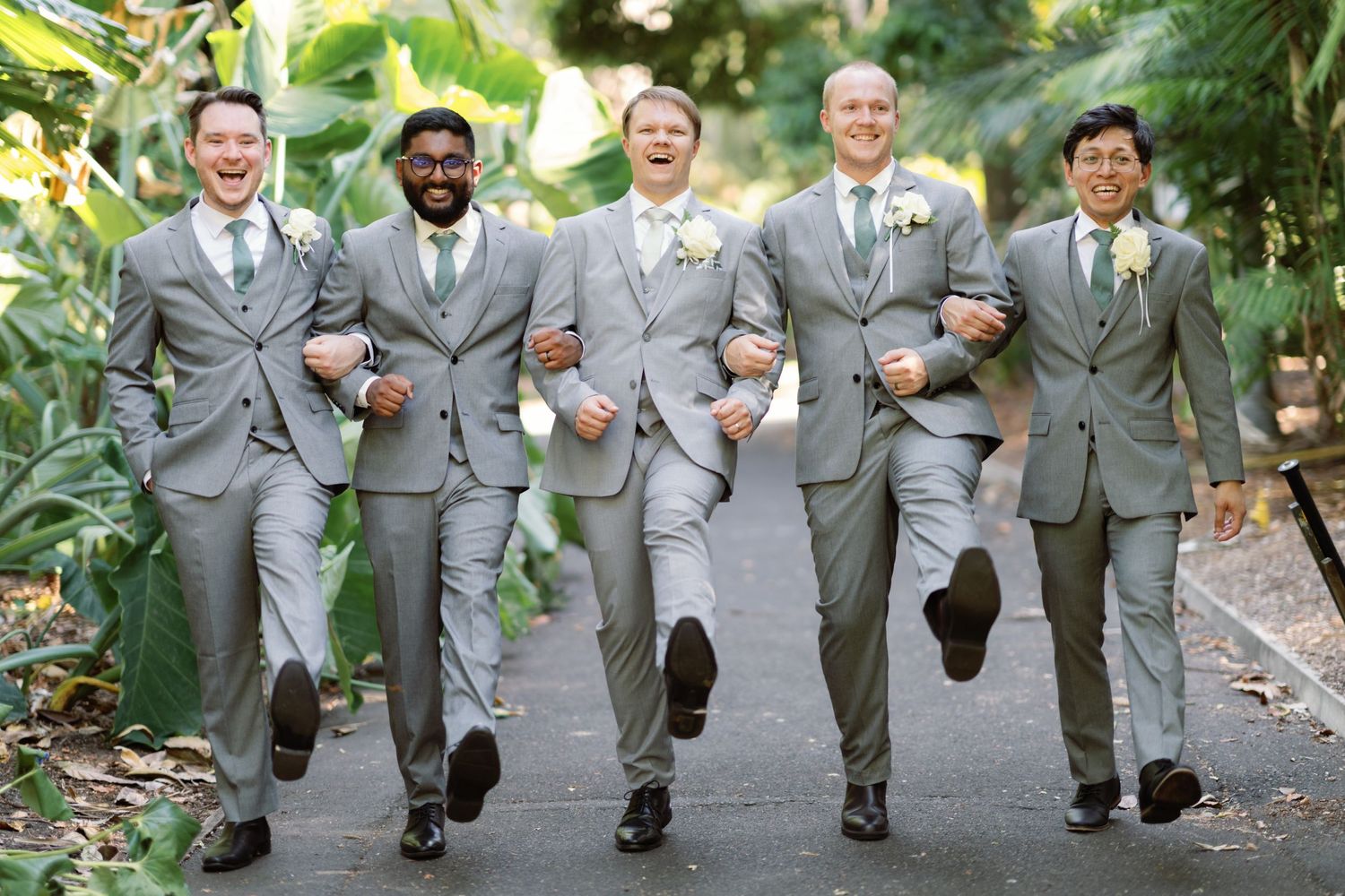 Groomsmen in matching gray suits jump and pose playfully on a paved pathway.