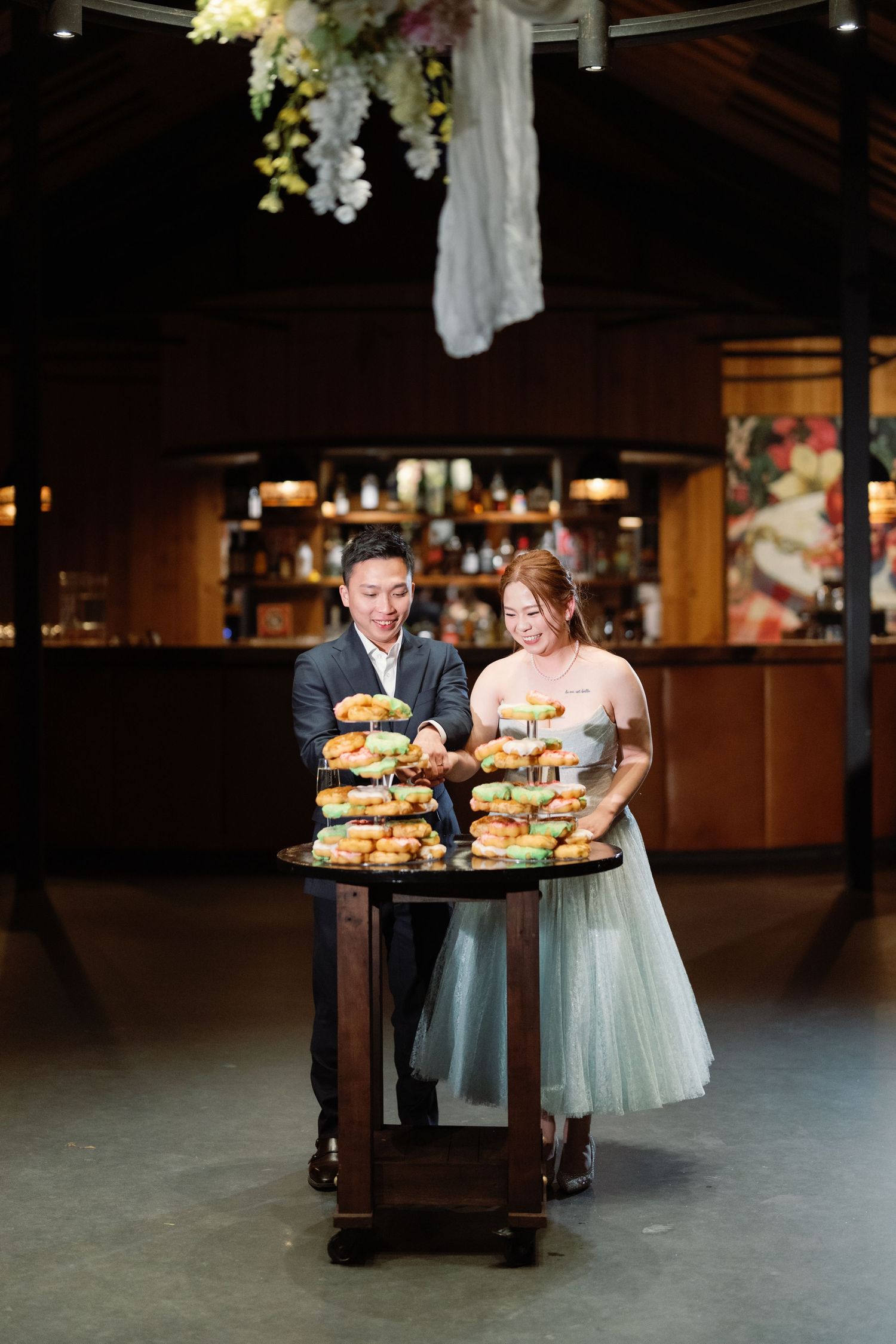A couple stands with a wooden cart displaying multiple tiers of gourmet burgers at an indoor reception venue.