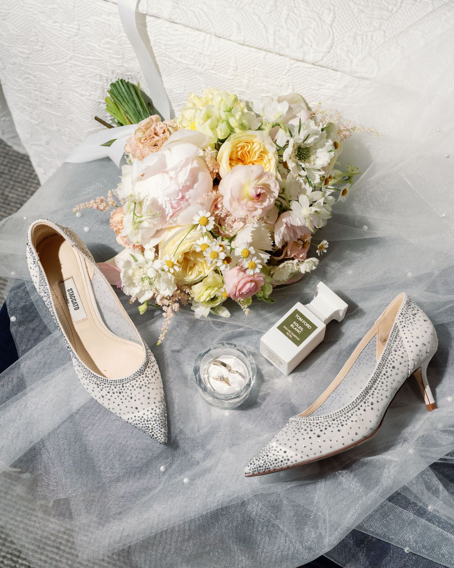 Elegant bridal accessories including sparkly heels, perfume bottle and pastel floral bouquet arranged on tulle fabric.