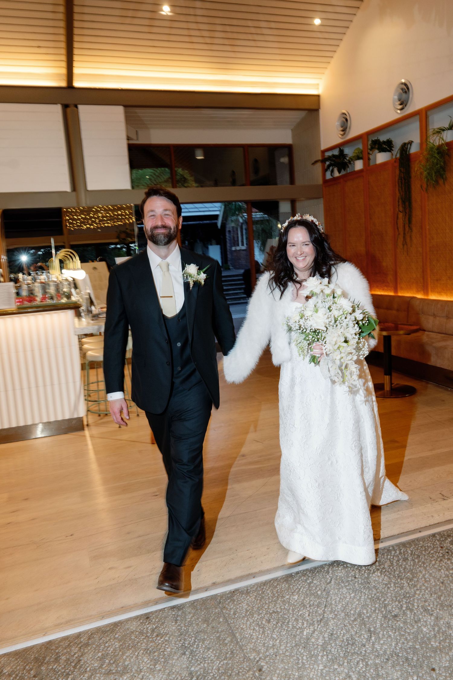 Smiling bride and groom make grand entrance into warmly lit wedding reception venue holding hands.