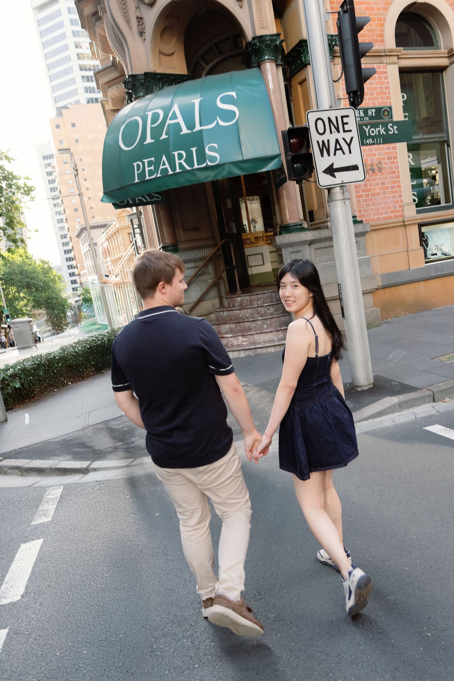 A couple in dark clothing walks down a city street past a storefront with green signage.