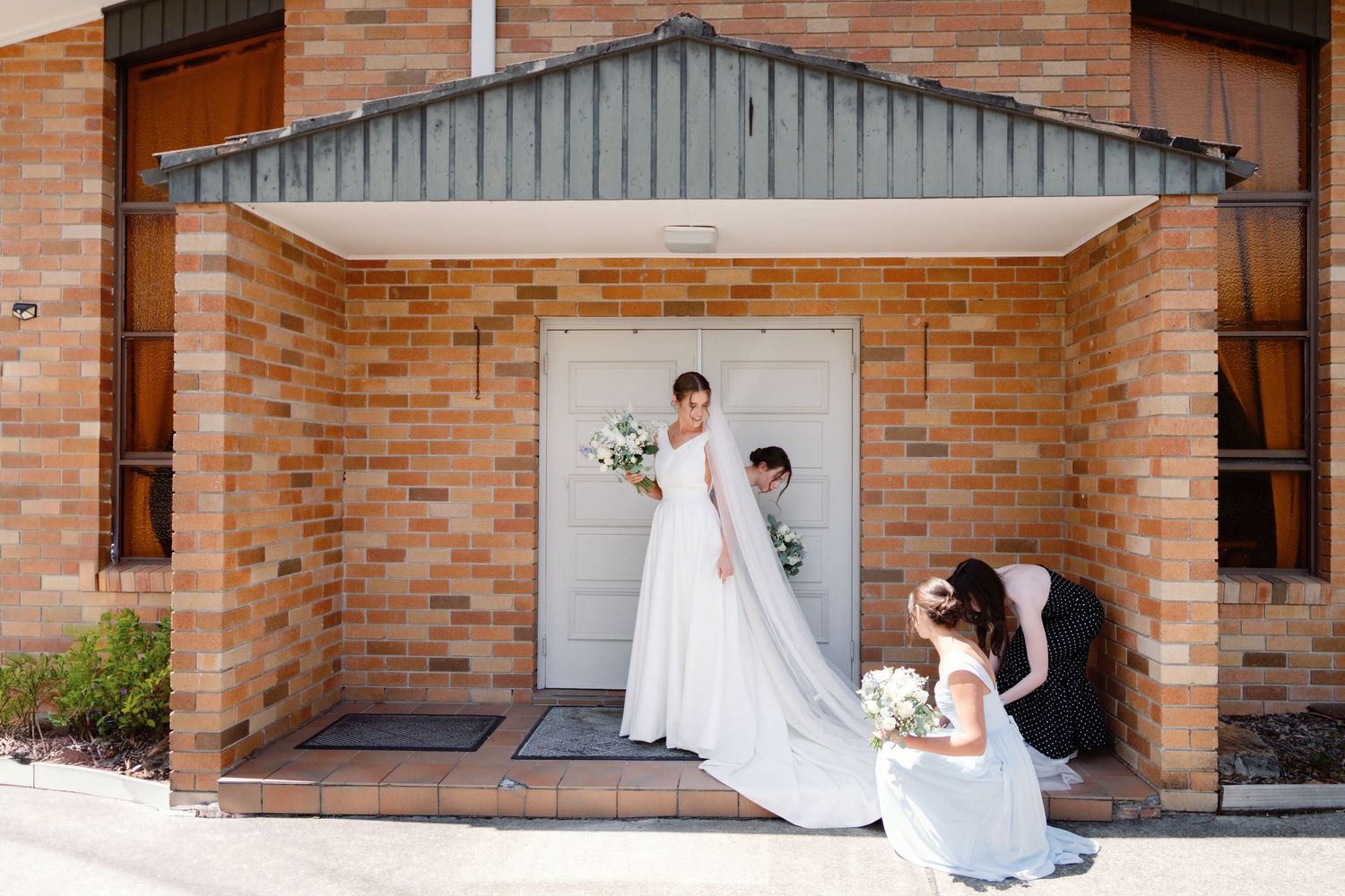 Wedding party helps adjust a bride's flowing white gown and veil outside a brick church entrance.