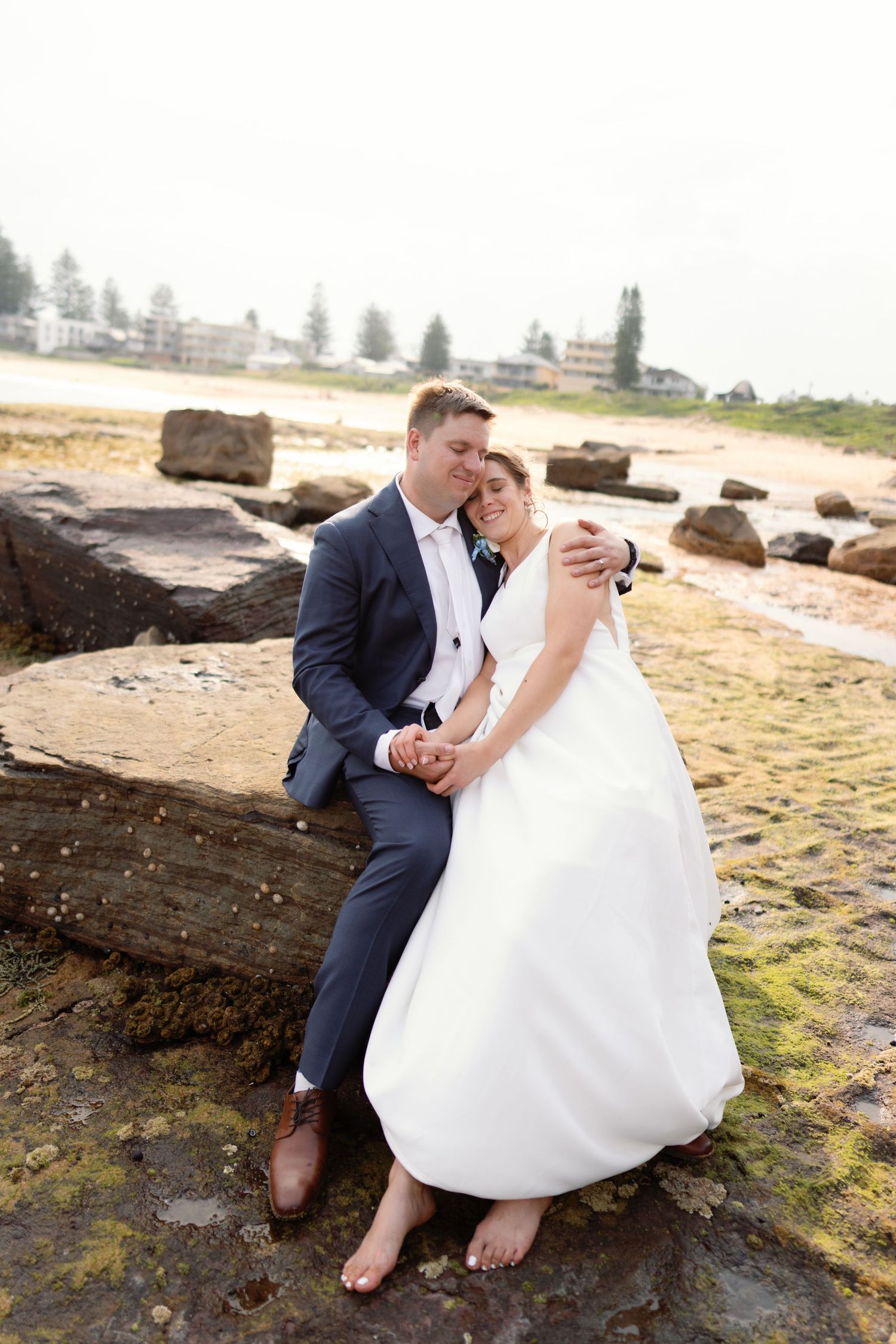Romantic wedding couple sitting on rocky beach during sunset photoshoot.