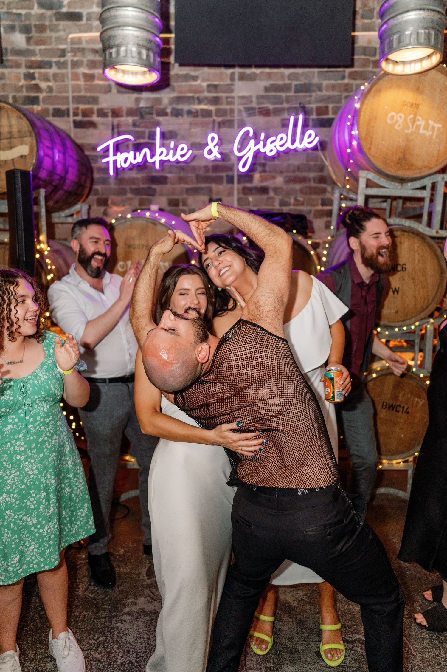 Party guests dancing and celebrating under a purple neon sign at a rustic venue with wooden barrels in the background.