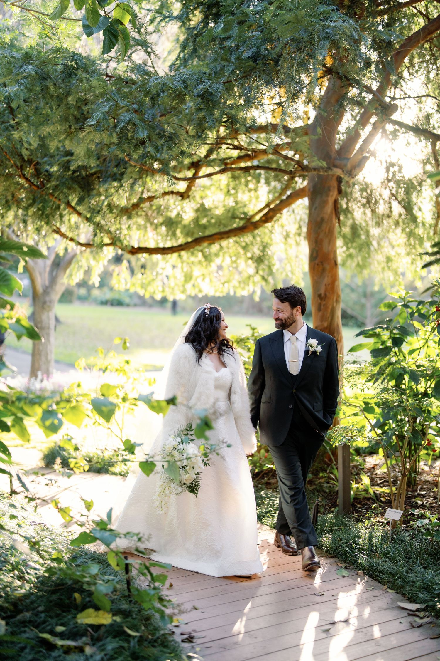 A couple shares a romantic moment under sunlit trees on a garden path during their wedding celebration.