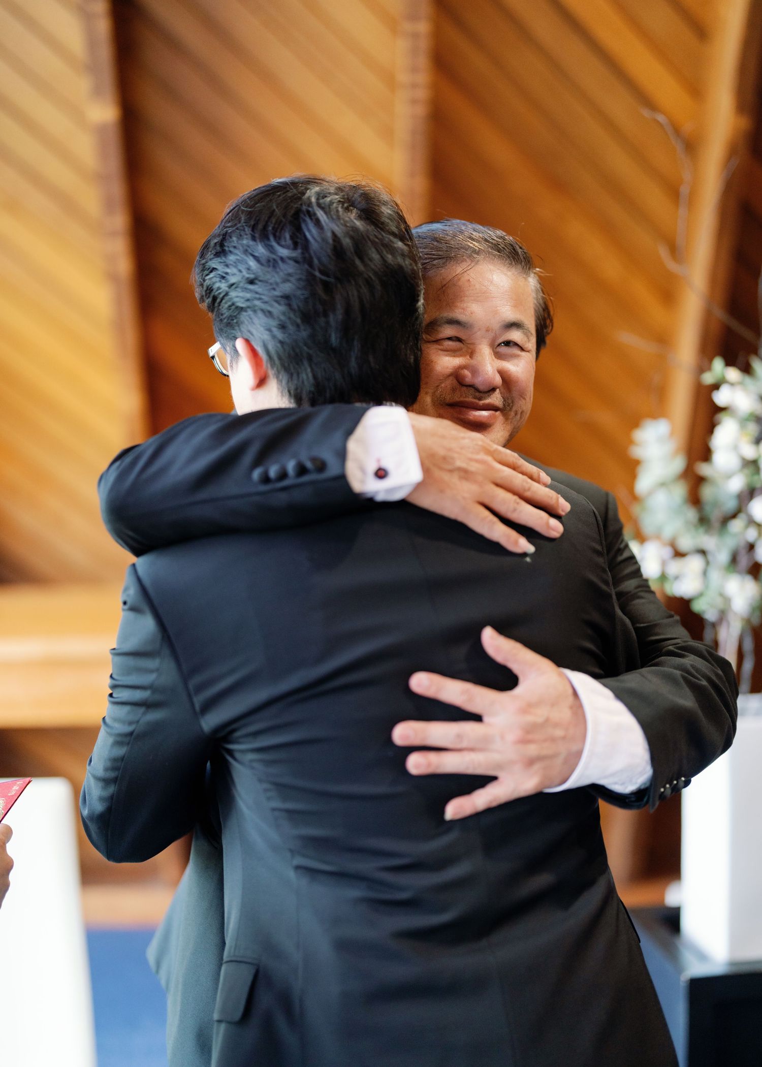 Two men in dark suits share a warm embrace during a wedding celebration in a wooden interior venue.