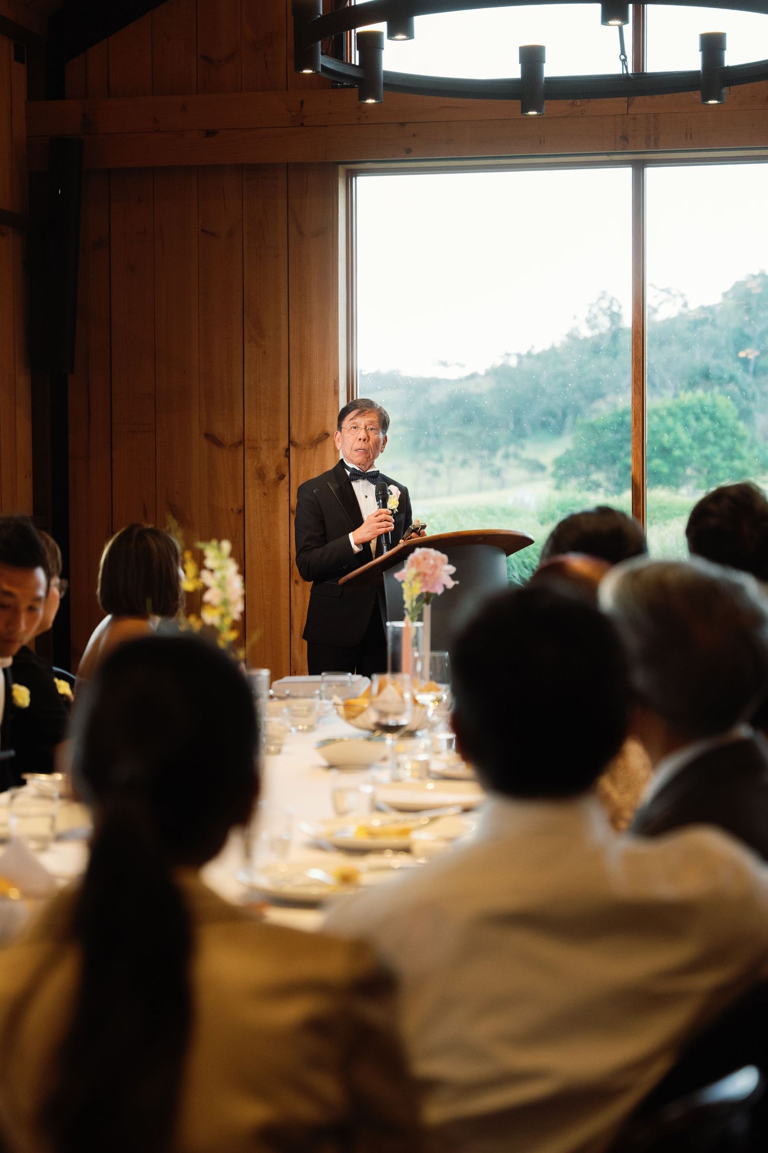 Speaker addresses wedding guests in wood-paneled reception room with large windows.