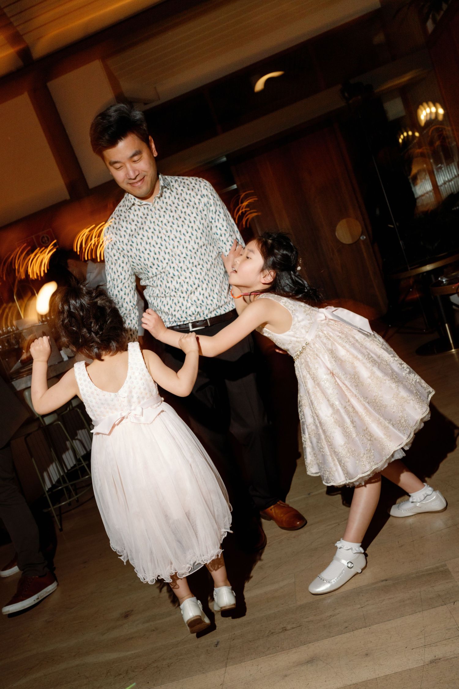 A man dances joyfully with two young girls in white dresses at a warm-lit wedding reception.