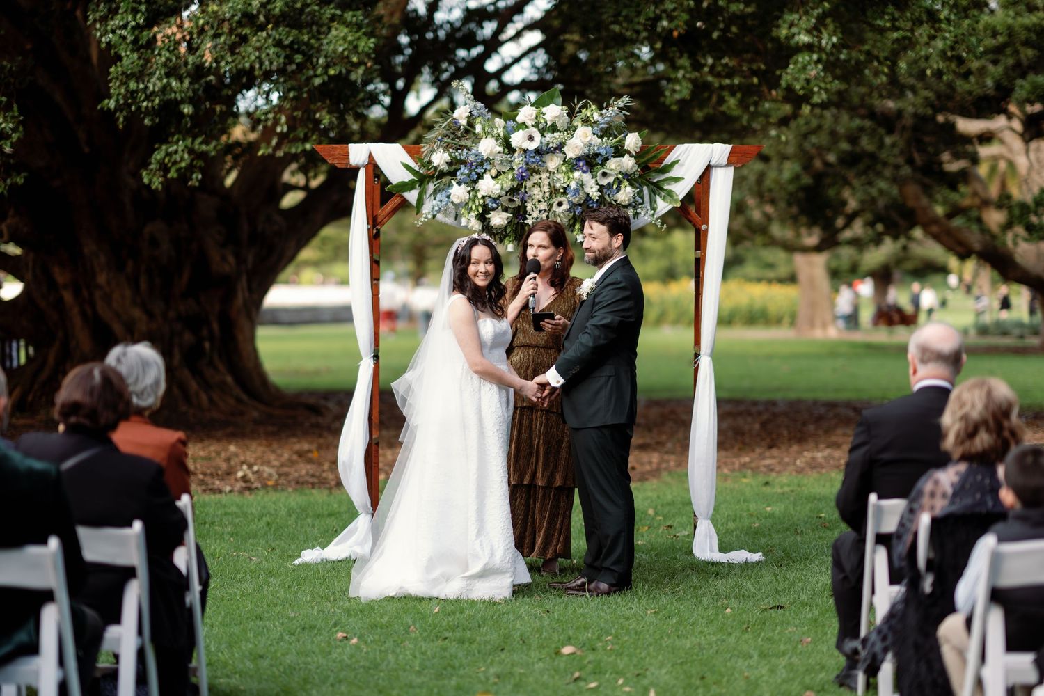 Outdoor wedding ceremony under floral arch with bride, groom, and officiant on green lawn beside large tree.