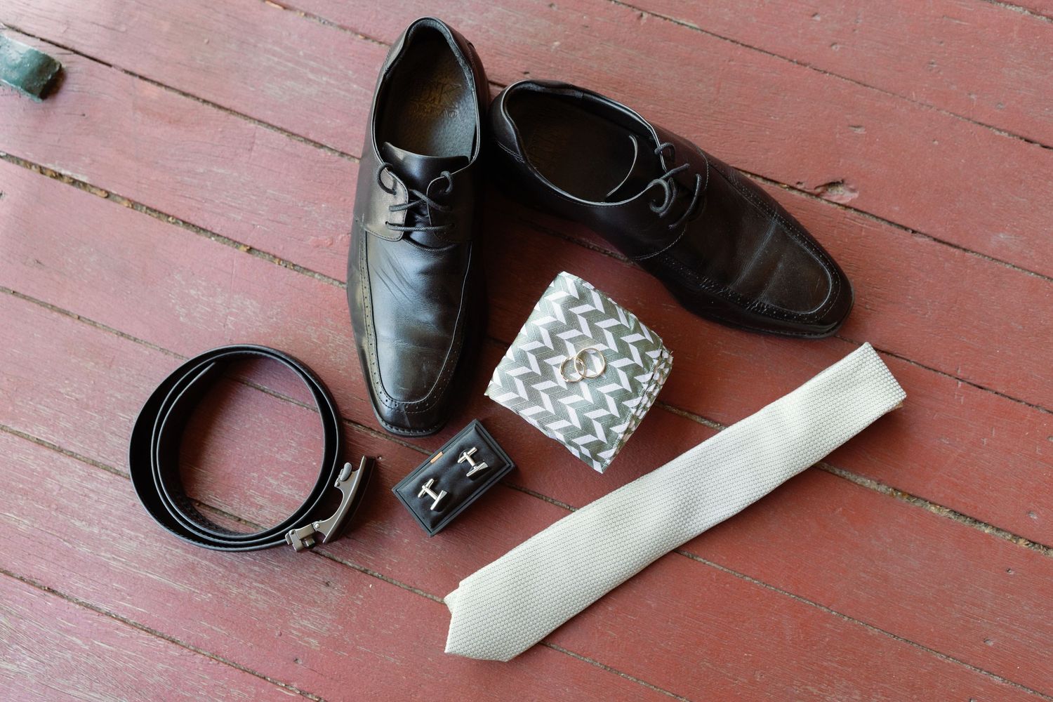 Leather dress shoes and accessories including a belt arranged on a rustic pink wooden floor.